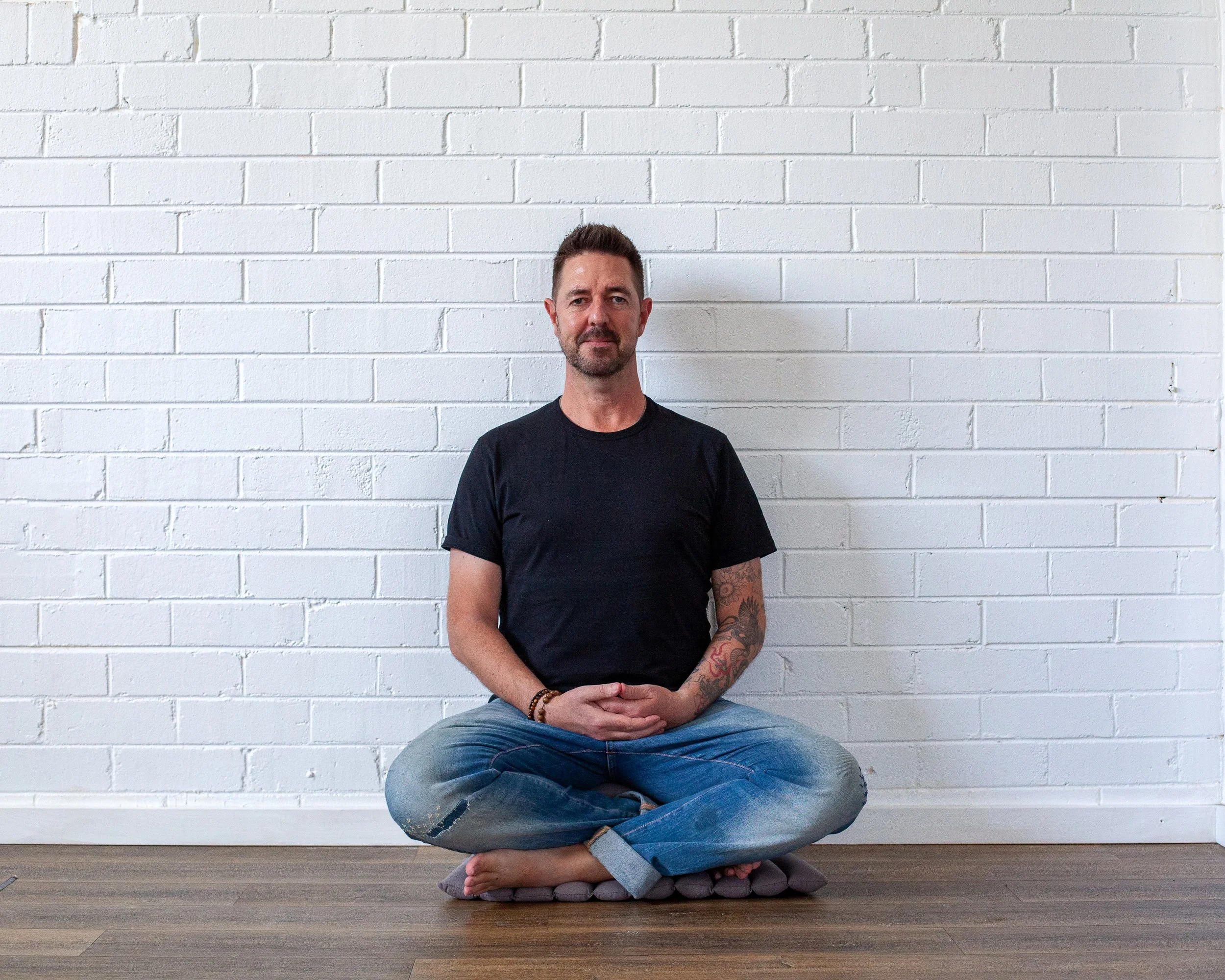A man with a beard and tattoos sitting cross-legged on a cushion in front of a white brick wall.