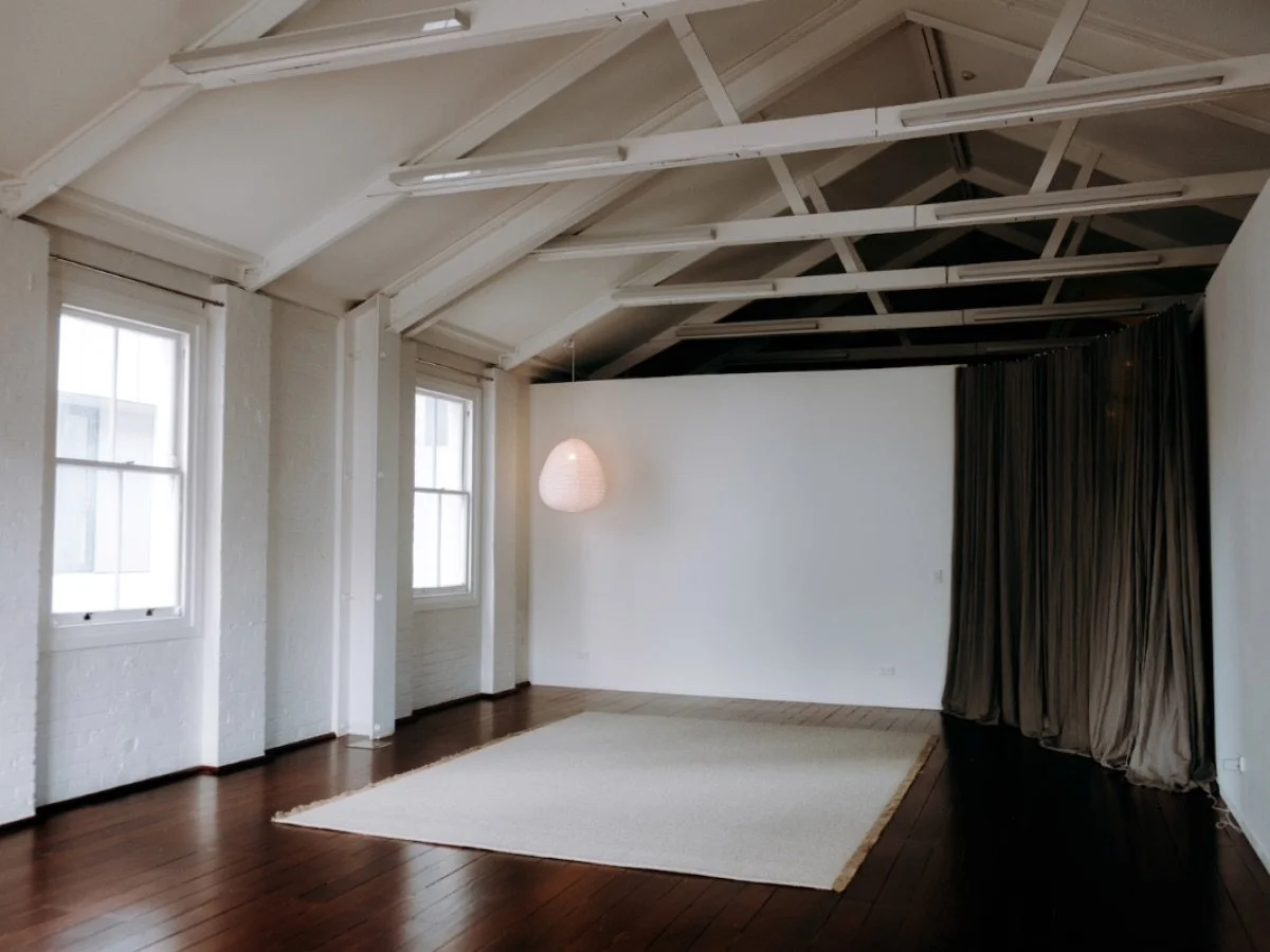 Spacious room with wooden floors, white walls, a hanging pendant light, and large windows. Features exposed ceiling beams and a white rug on the floor.