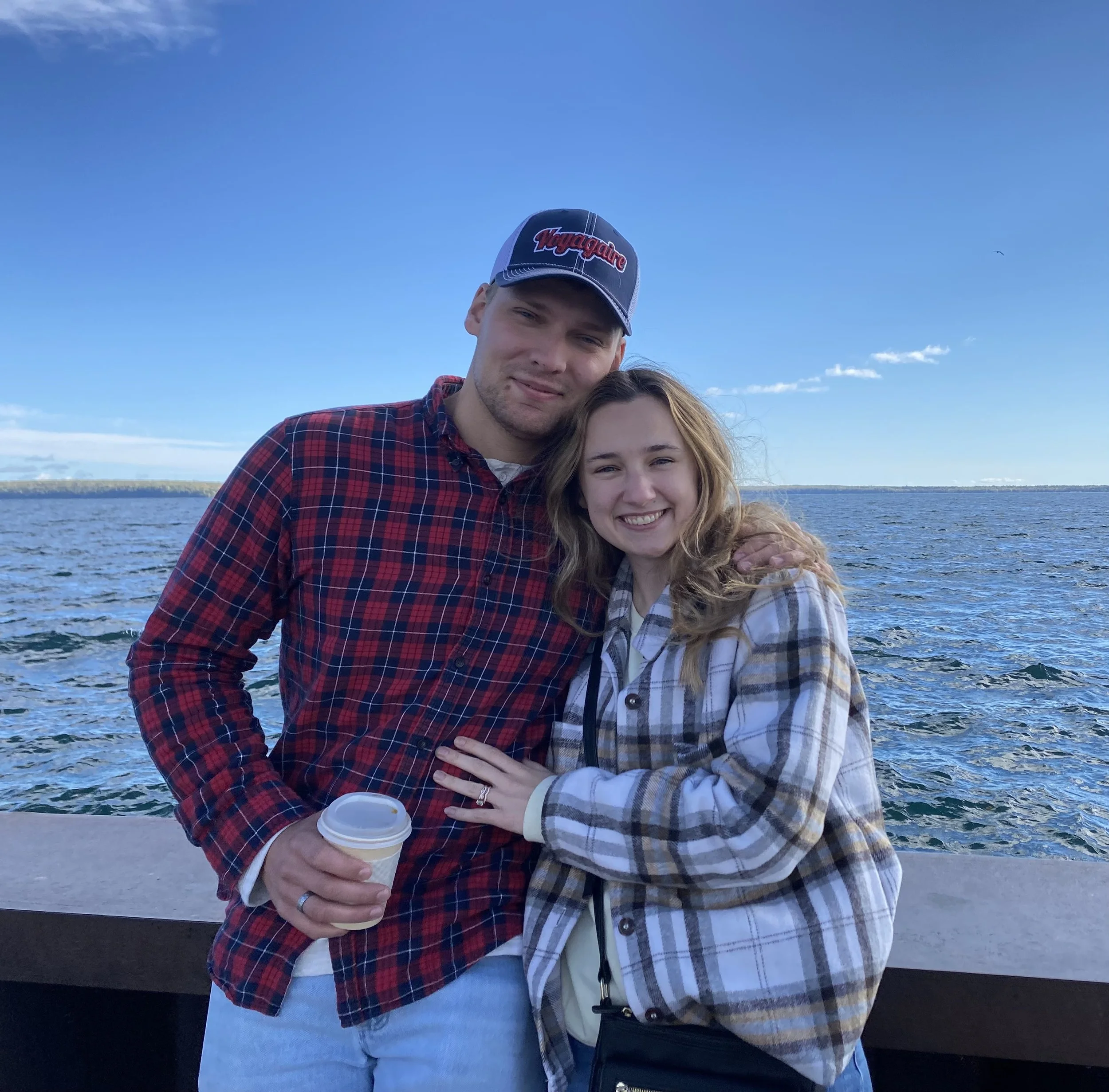 Smiling couple posing by the ocean, wearing plaid shirts, holding a coffee cup, with blue sky in the background.