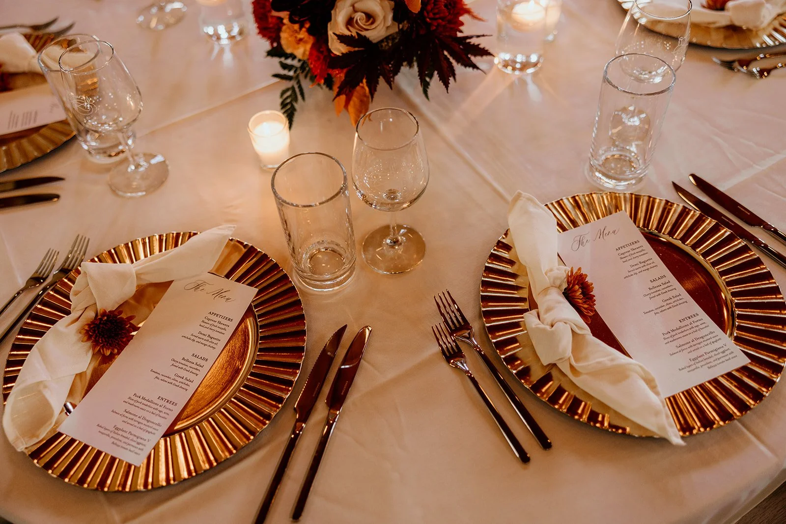 Elegant table setting with gold chargers, menus, white napkins, glassware, cutlery, and flower centerpiece.