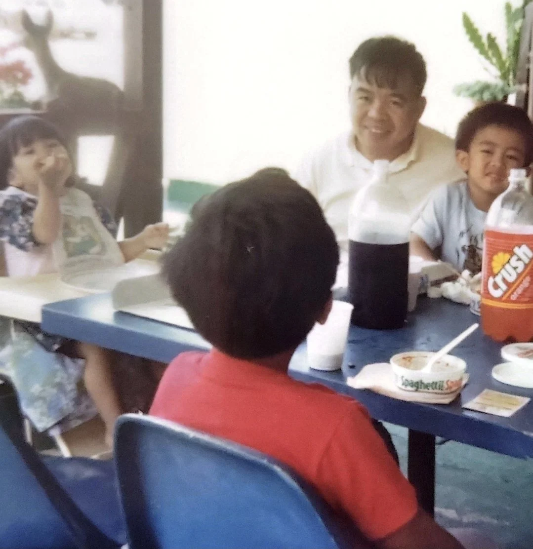 A old 90s era picture of a family, group of kids and an adult, sitting at a table eating spaghetti with drinks and snacks, smiling and enjoying their time together.