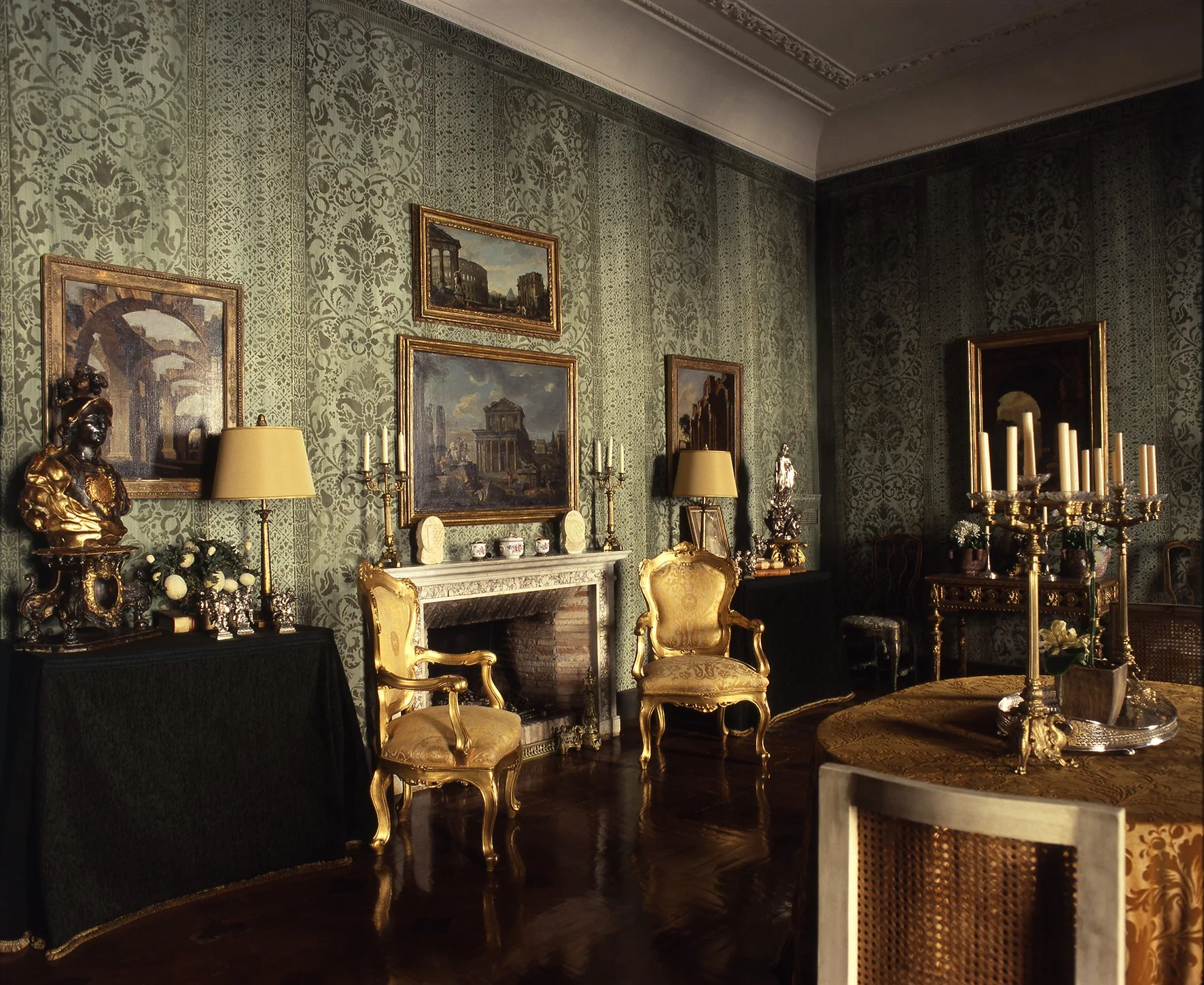 Luxurious vintage living room with ornate green wallpaper, gold chairs, framed artwork, a marble fireplace, and a table with tall candle holders.