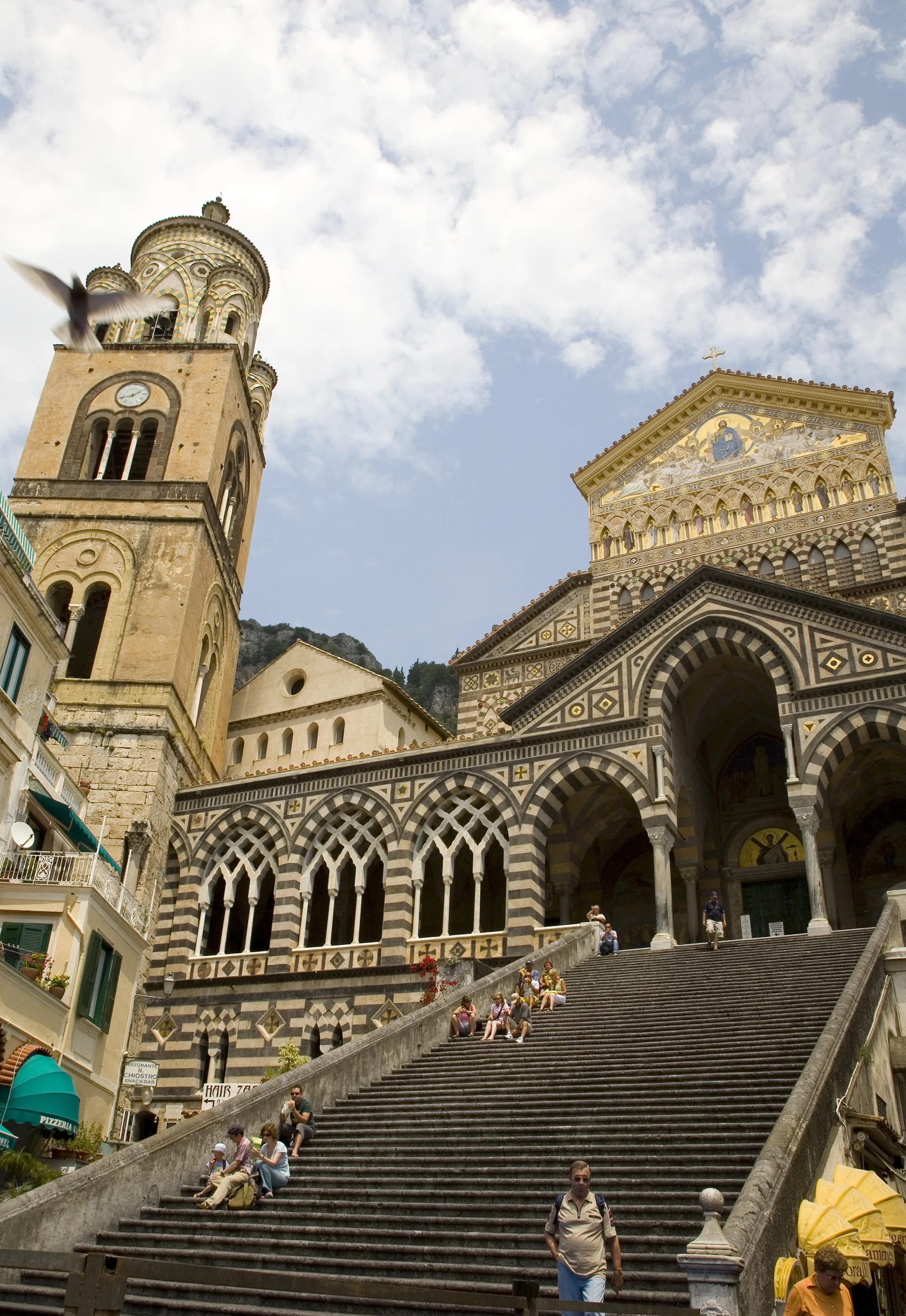 People sitting on a large staircase outside a historical church with striped black and white arches, a clock tower, and a golden tiled roof with an icon of Jesus above the entrance, under a partly cloudy sky.