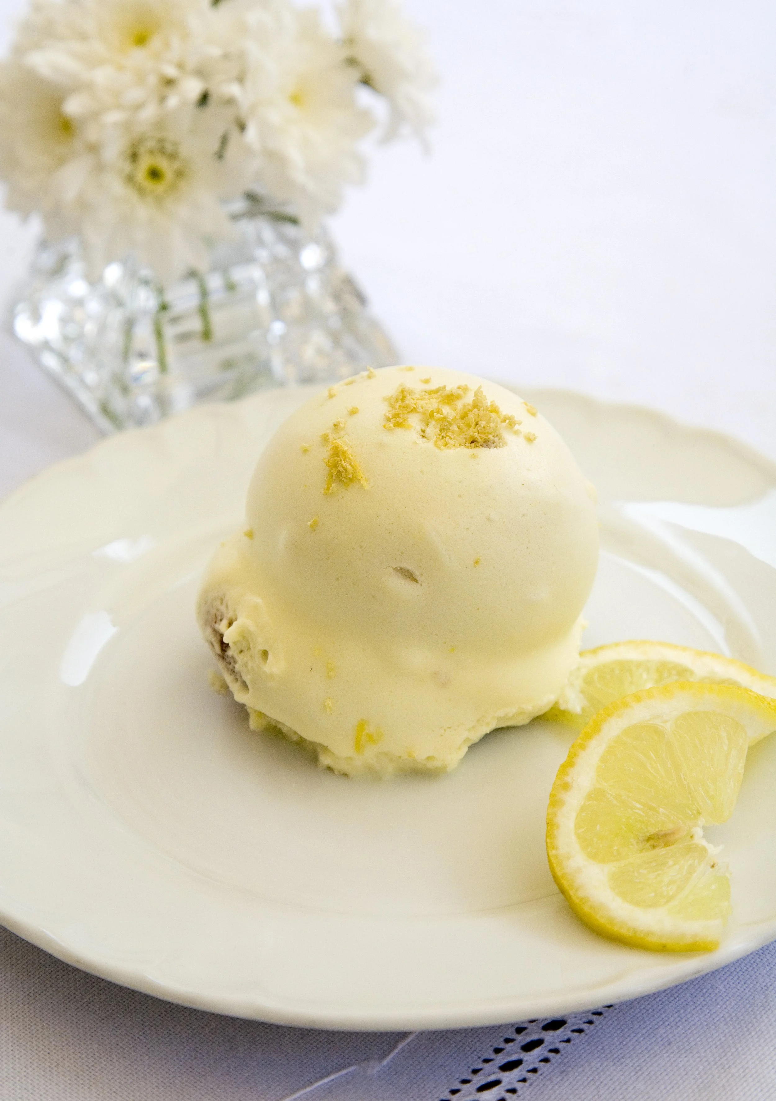 Scoops of vanilla ice cream garnished with lemon zest, served with lemon wedges on a white plate, with a blurred white flower arrangement in the background.