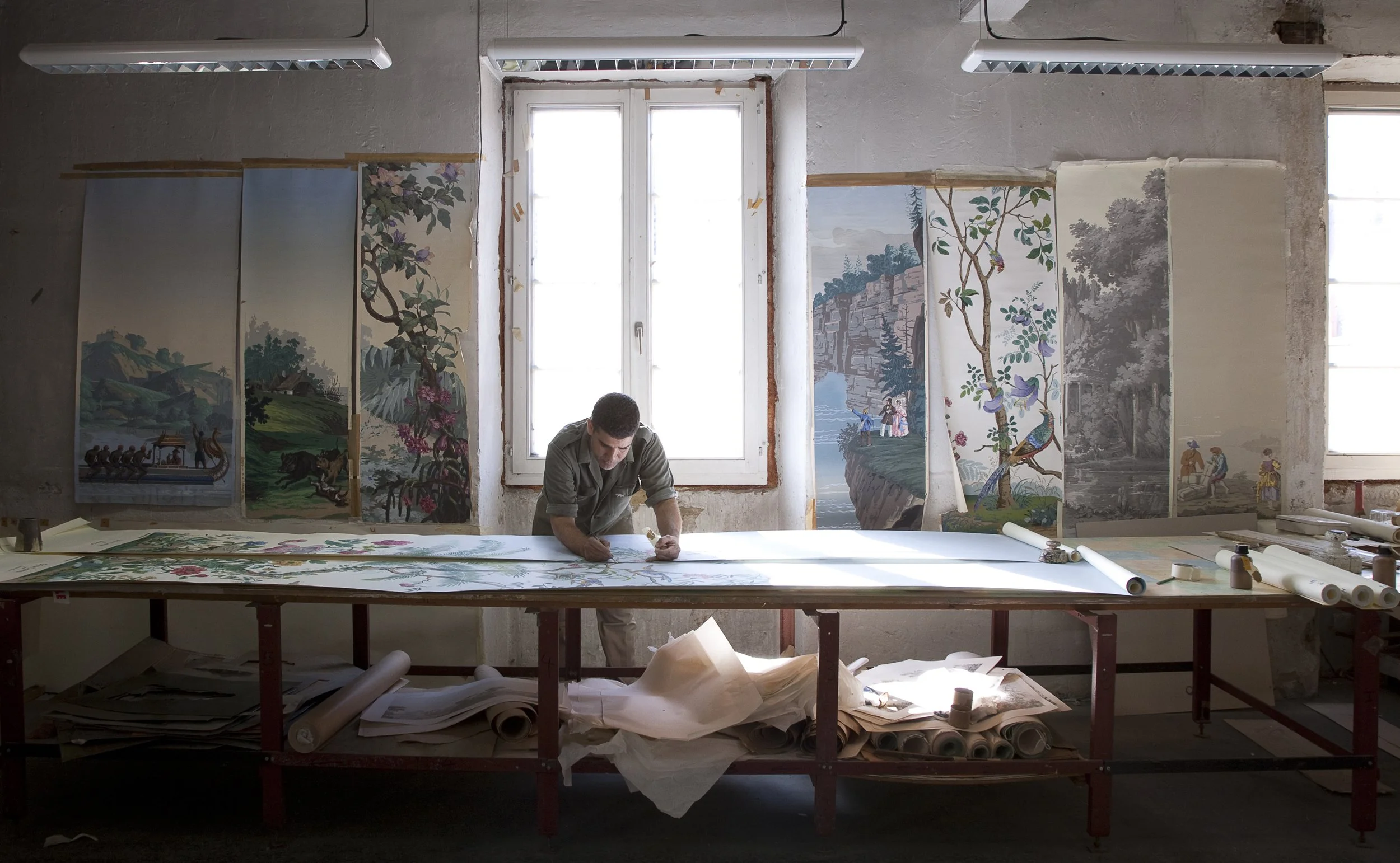An artist working on a large painting in a studio with several traditional-style landscape and nature paintings hanging on the wall behind him, illuminated by natural light from two windows.