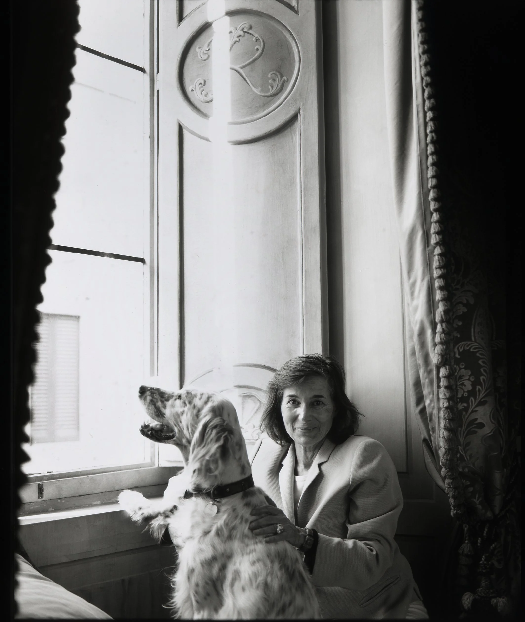 A woman sitting by a window with a dog, looking outside. The woman is smiling and wearing a light-colored blazer. The dog, an Irish Setter, is sitting on her lap with its paws on the window ledge, looking outside.