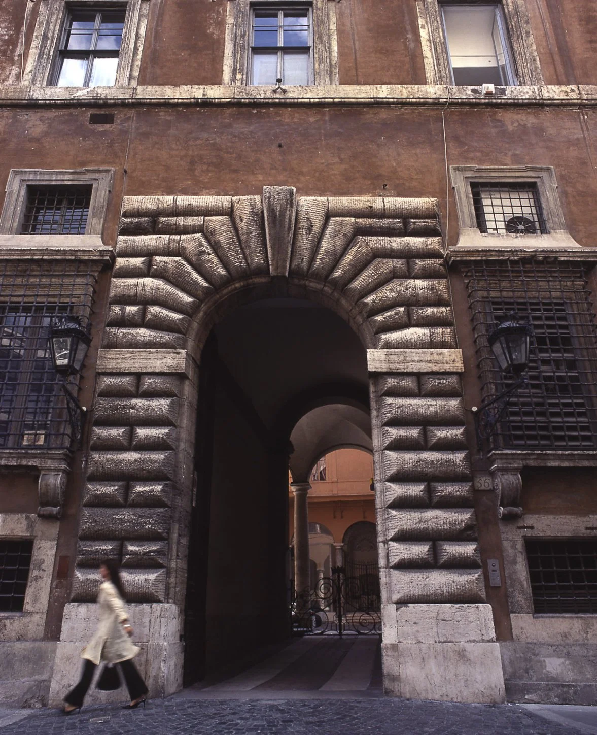 View of a historic building with a large arched entryway, stonework detail around the arch, and barred windows. A woman is walking by in front of the building.