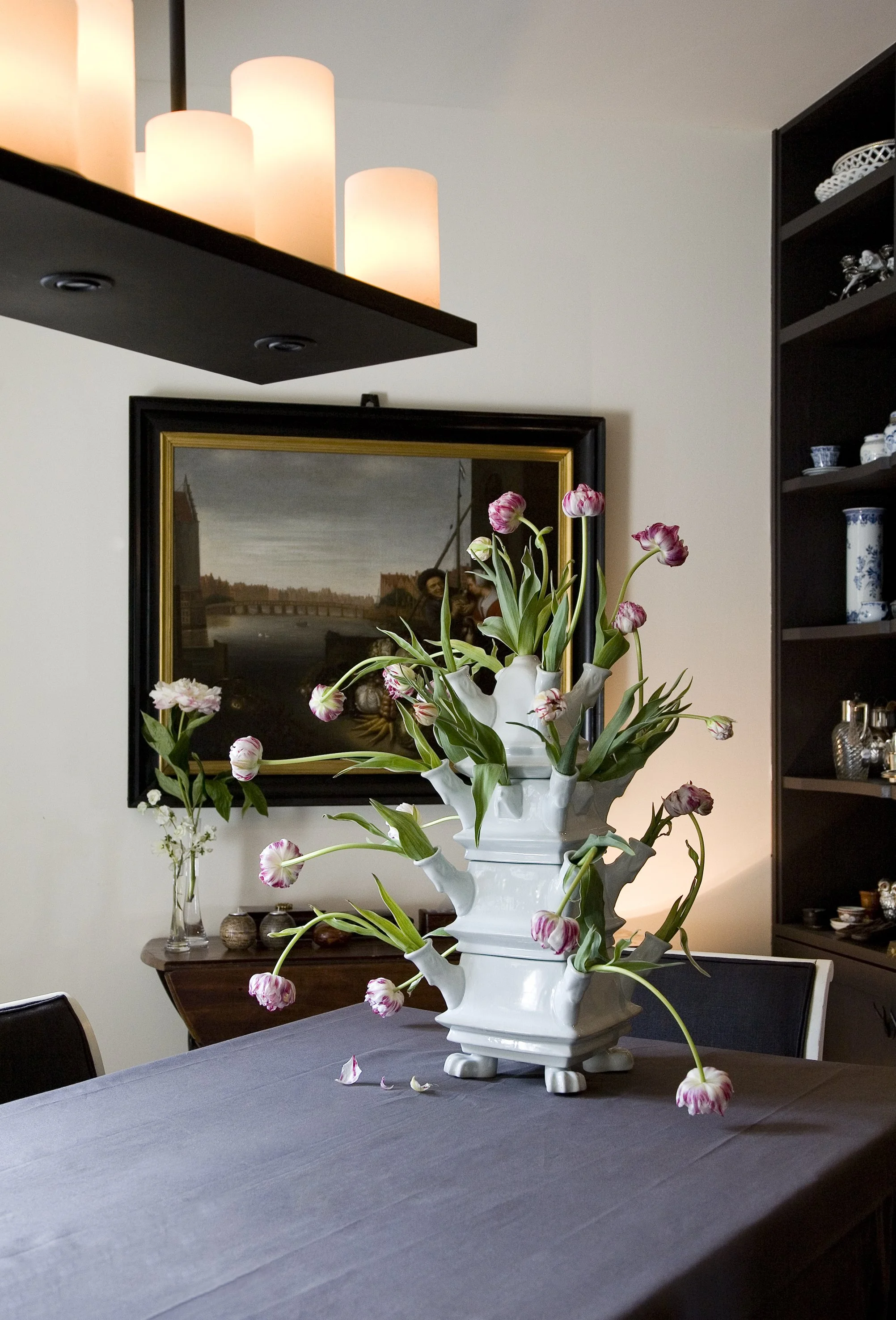 A large white ceramic vase with pink and white tulips on a dining table. The background includes a dark wooden shelf with decorative items and a framed painting on the wall.