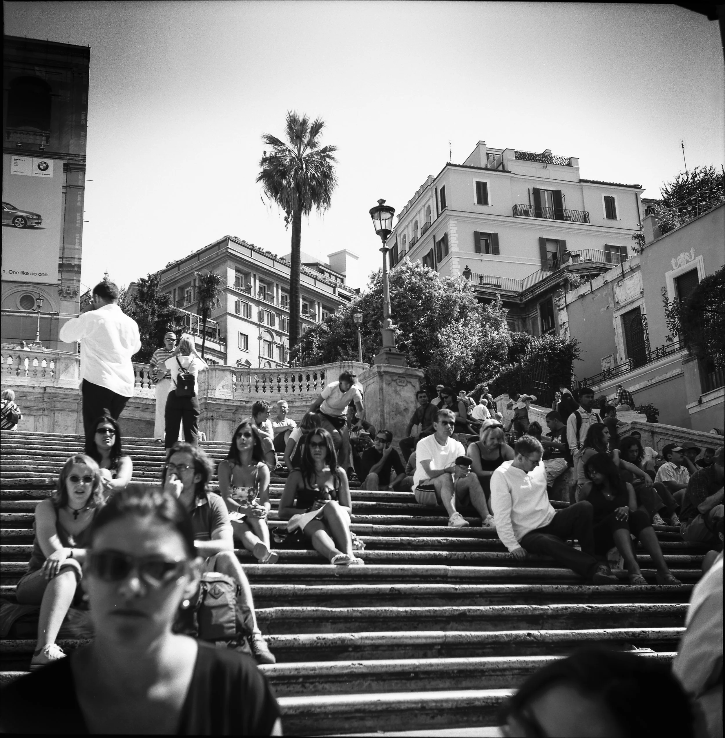 People sitting and standing on an outdoor staircase in an urban area with buildings, trees, and a palm tree in the background
