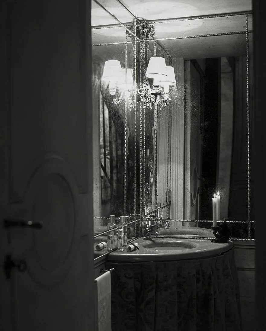 A black-and-white photograph of a vintage bathroom with a round sink, bottles, a candle, and a chandelier reflected in a mirror with hanging beads, creating a moody atmosphere.