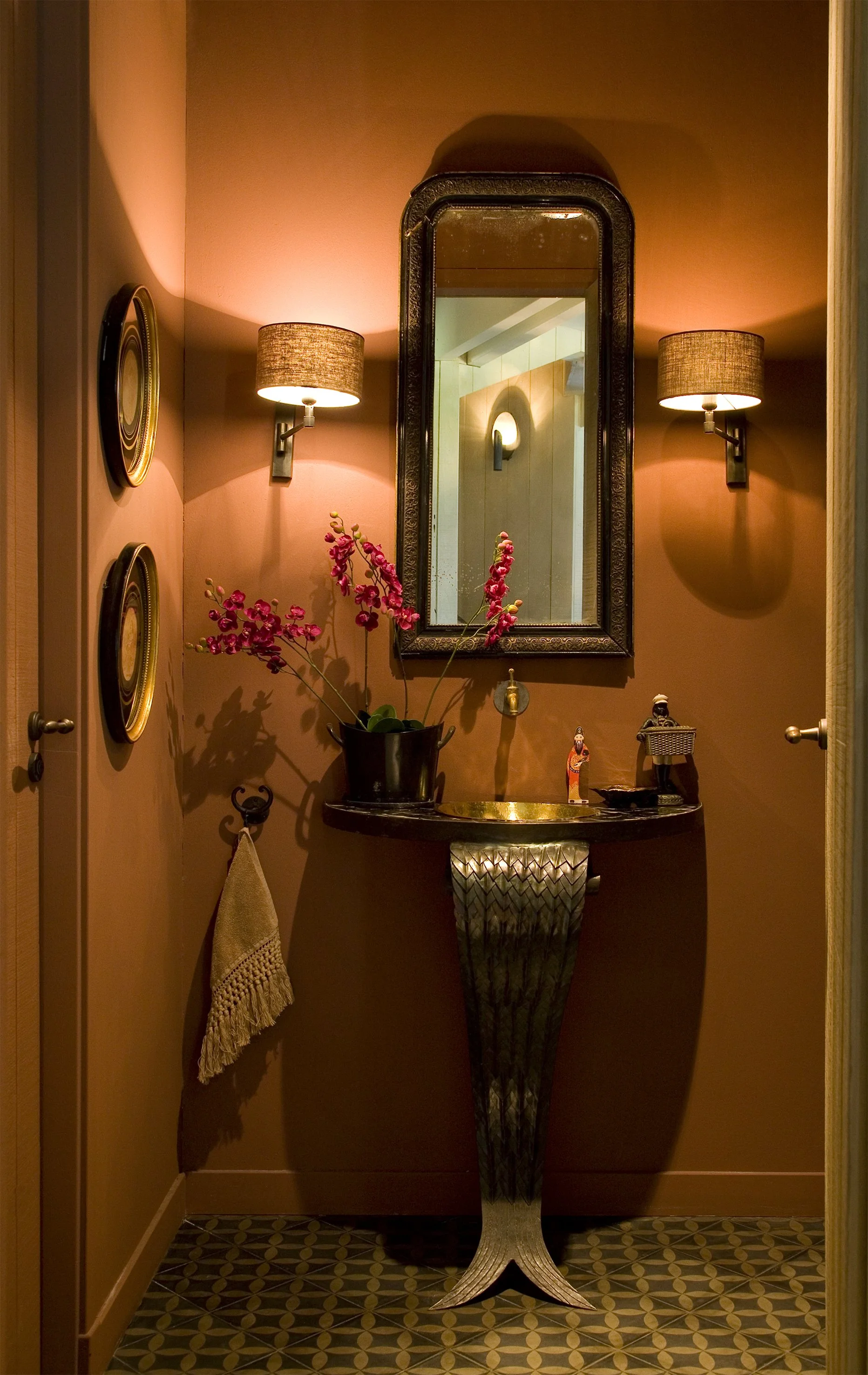 A small bathroom with peach-colored walls, a decorative mirror, two wall-mounted lamps, a black vase with pink orchids, and a vintage-style sink with a metallic finish and design. There are decorative plates on the wall and a towel hanging on a hook.