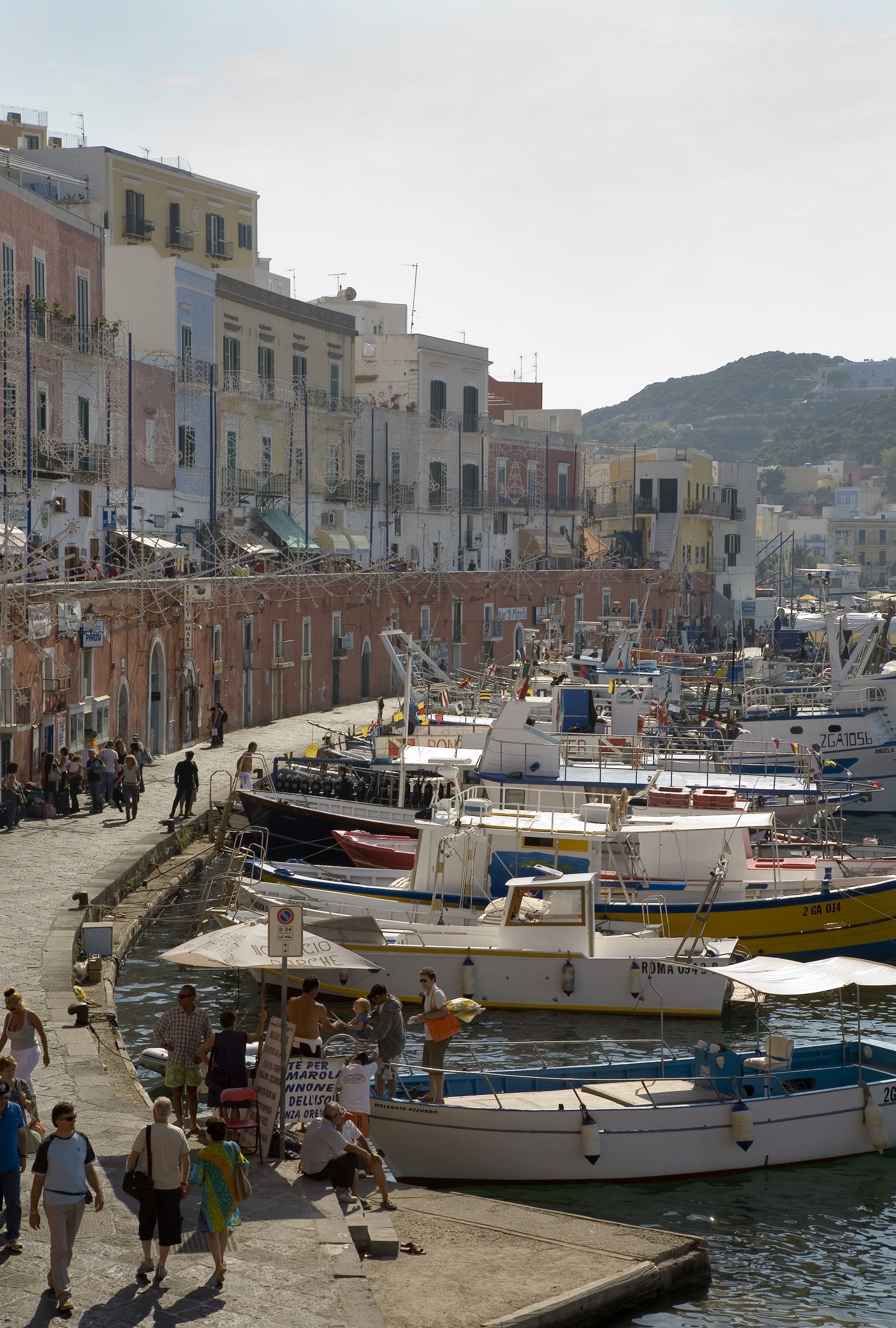 Colorful buildings lining a waterfront with boats docked along the pier, and people walking and sitting on the dock in a sunny coastal town.
