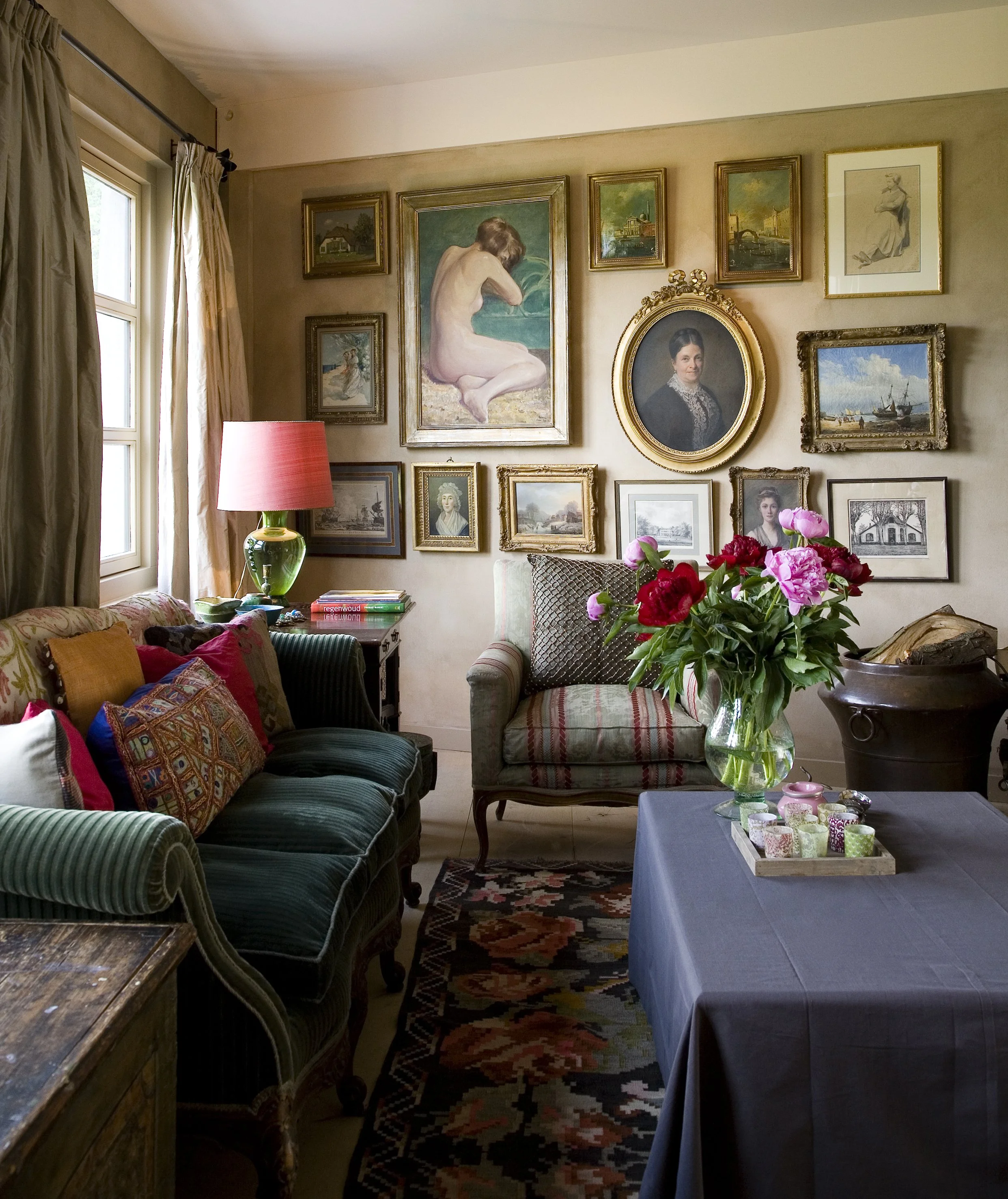 A cozy living room with a wall decorated with framed artwork and a portrait. There is a window with curtains on the left, a dark green sofa with colorful pillows, a small table with books, a side table with a pink lampshade, a vintage armchair with a cushion, a large vase with pink and red flowers, and a table covered with a blue cloth displaying candles.