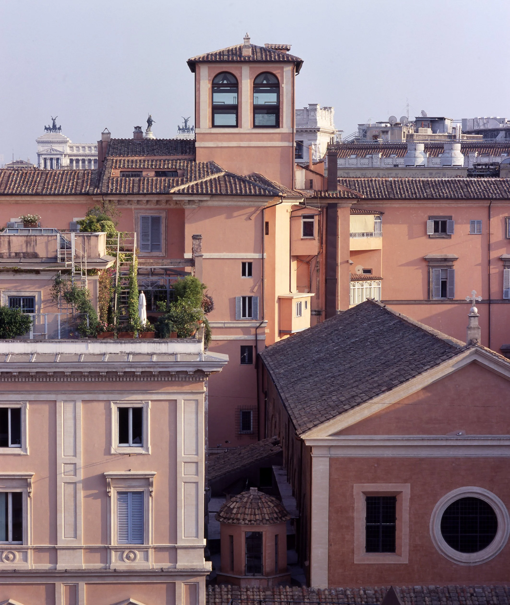 A cityscape of old European buildings with pink and beige facades, tiled roofs, and small windows, including a prominent building with a tower and large arched windows, against a cloudy sky.