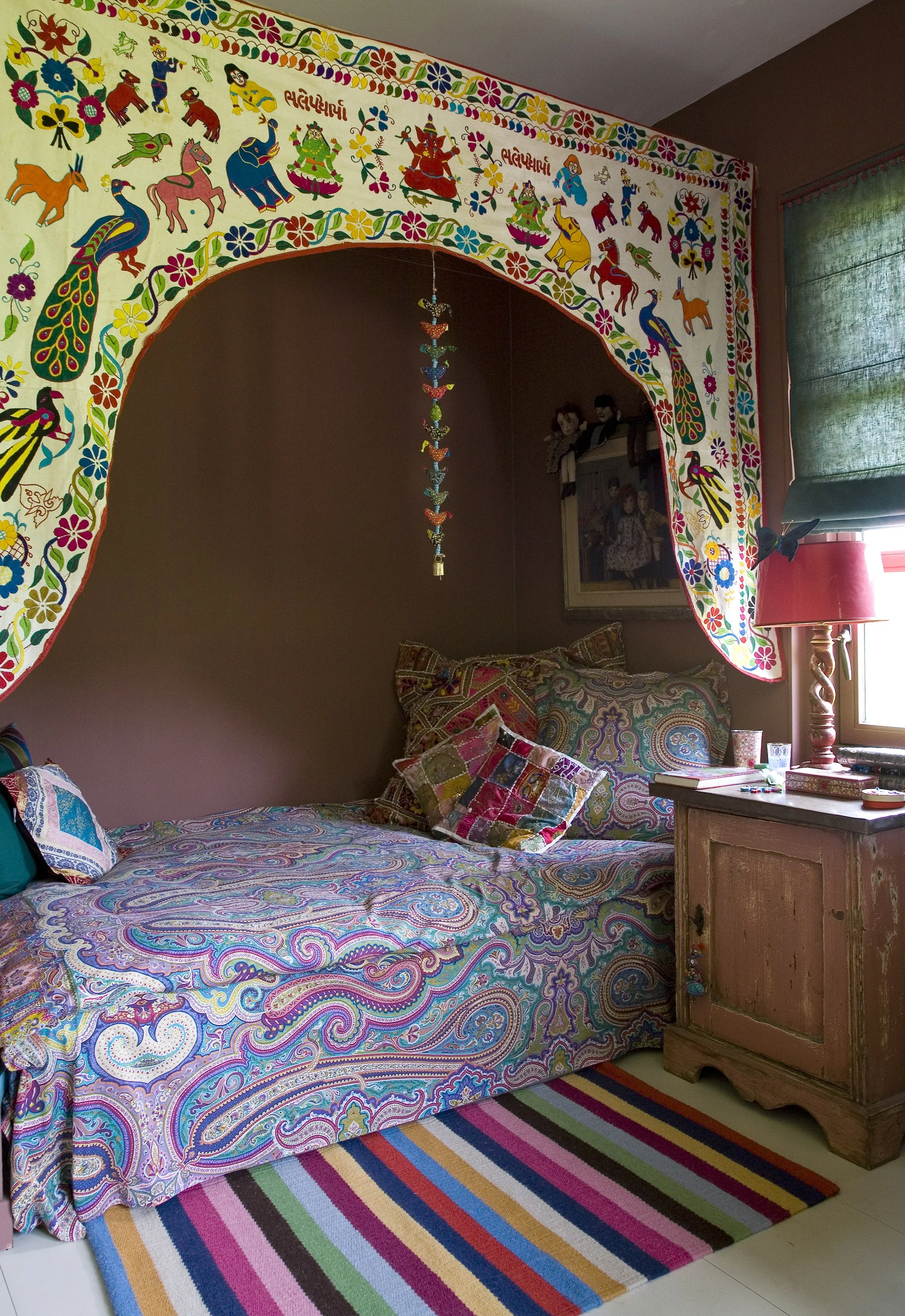 A bedroom decorated with colorful, paisley-patterned bedding and pillows, a wooden bedside table with a red lamp, and a gallery wall with dolls and framed photos. A striped rainbow rug is on the floor, and an ornate, horned arch with Indian-style artwork and embroidery adorns the top of the bed.