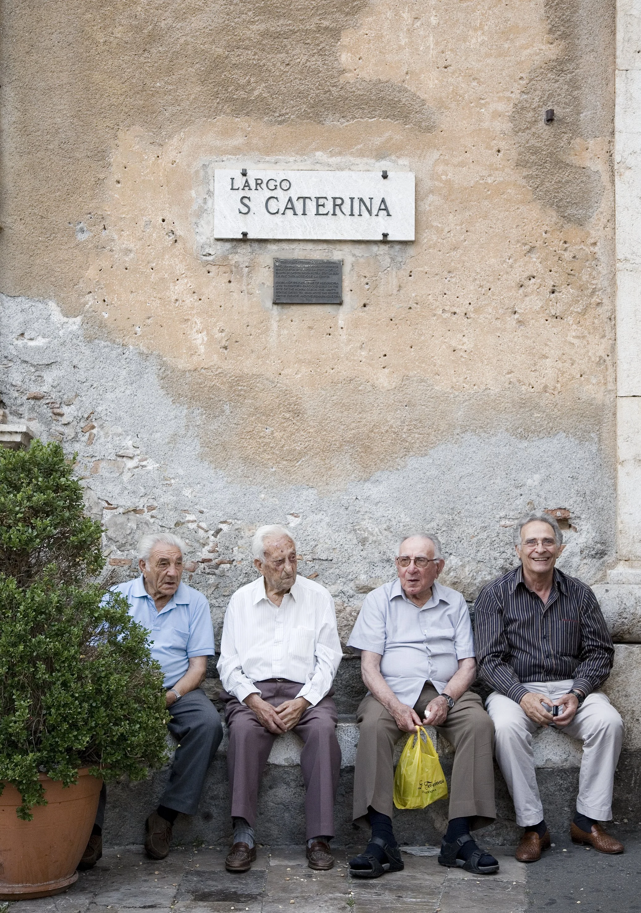 Four elderly men sit on a stone bench in front of a weathered beige wall with a street sign that reads "Largo S. Caterina." They are casually dressed and appear to be having a conversation.