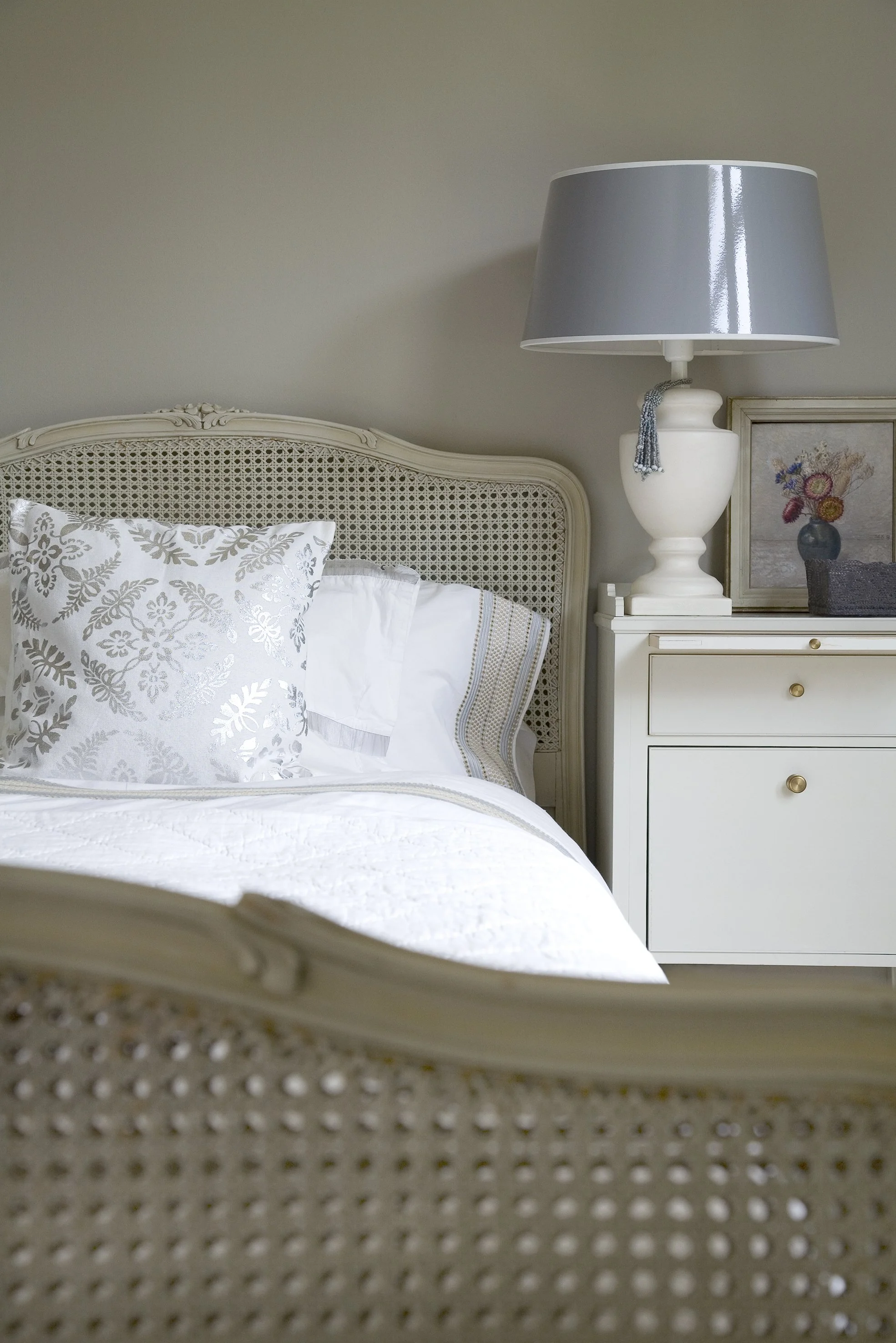 A neatly made bed with white and patterned pillows, a white headboard, and a white nightstand with a large gray lampshade and decorative items.