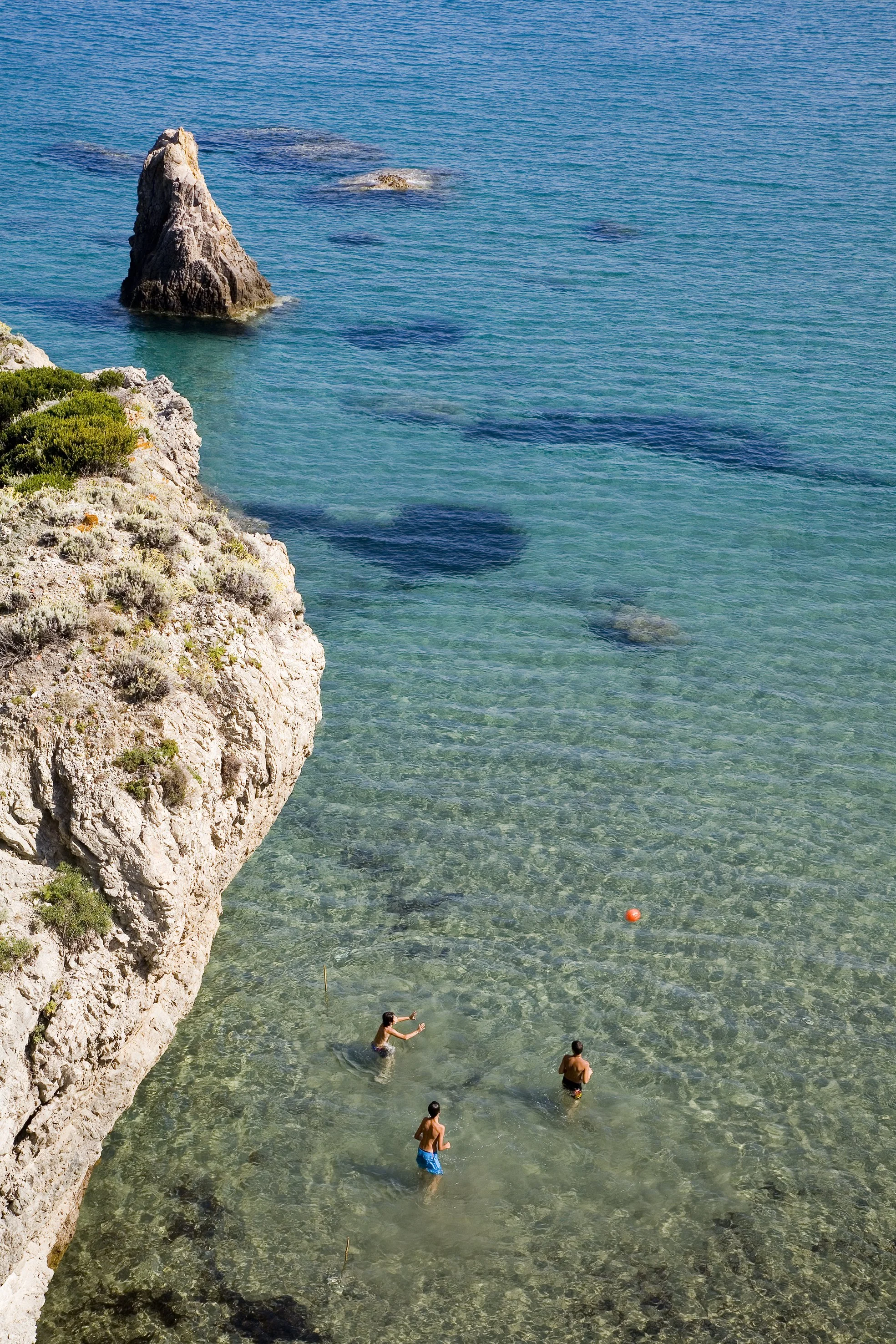 Three children playing in shallow, clear water near a rocky beach with a large rock formation in the background.