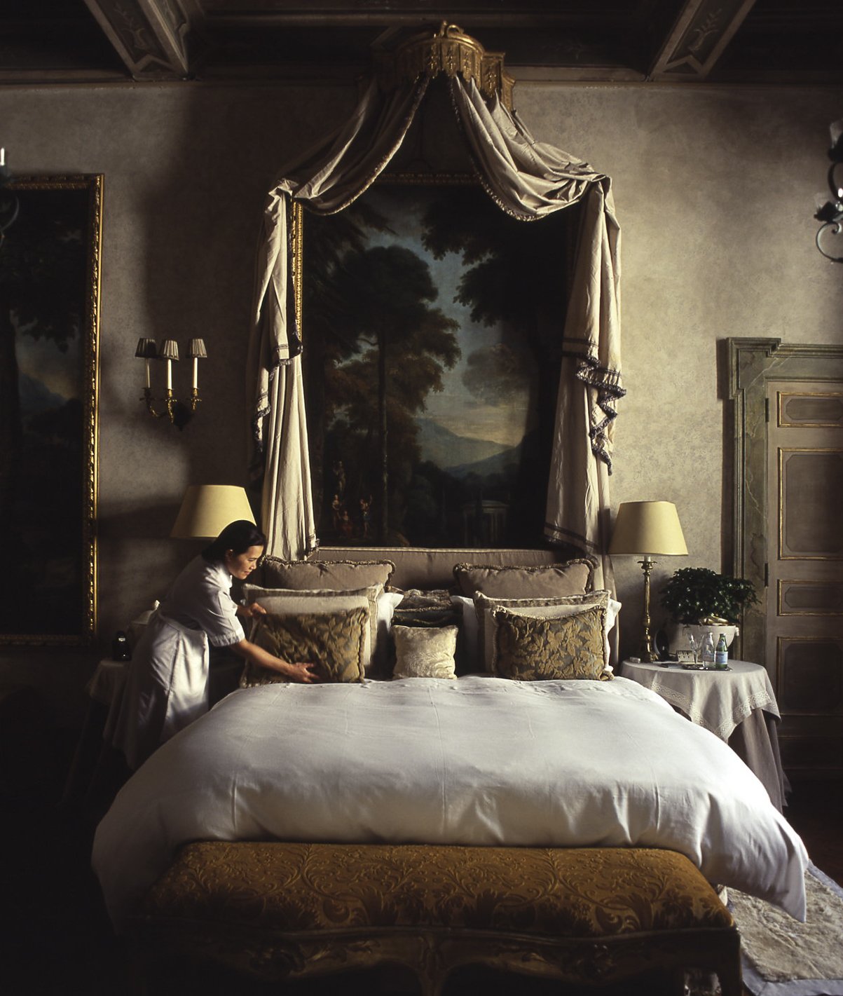 A woman making a bed in an elegant, vintage-style bedroom with a large bed, ornate pillows, a canopy with draped curtains, framed landscape paintings, and antique lamps.