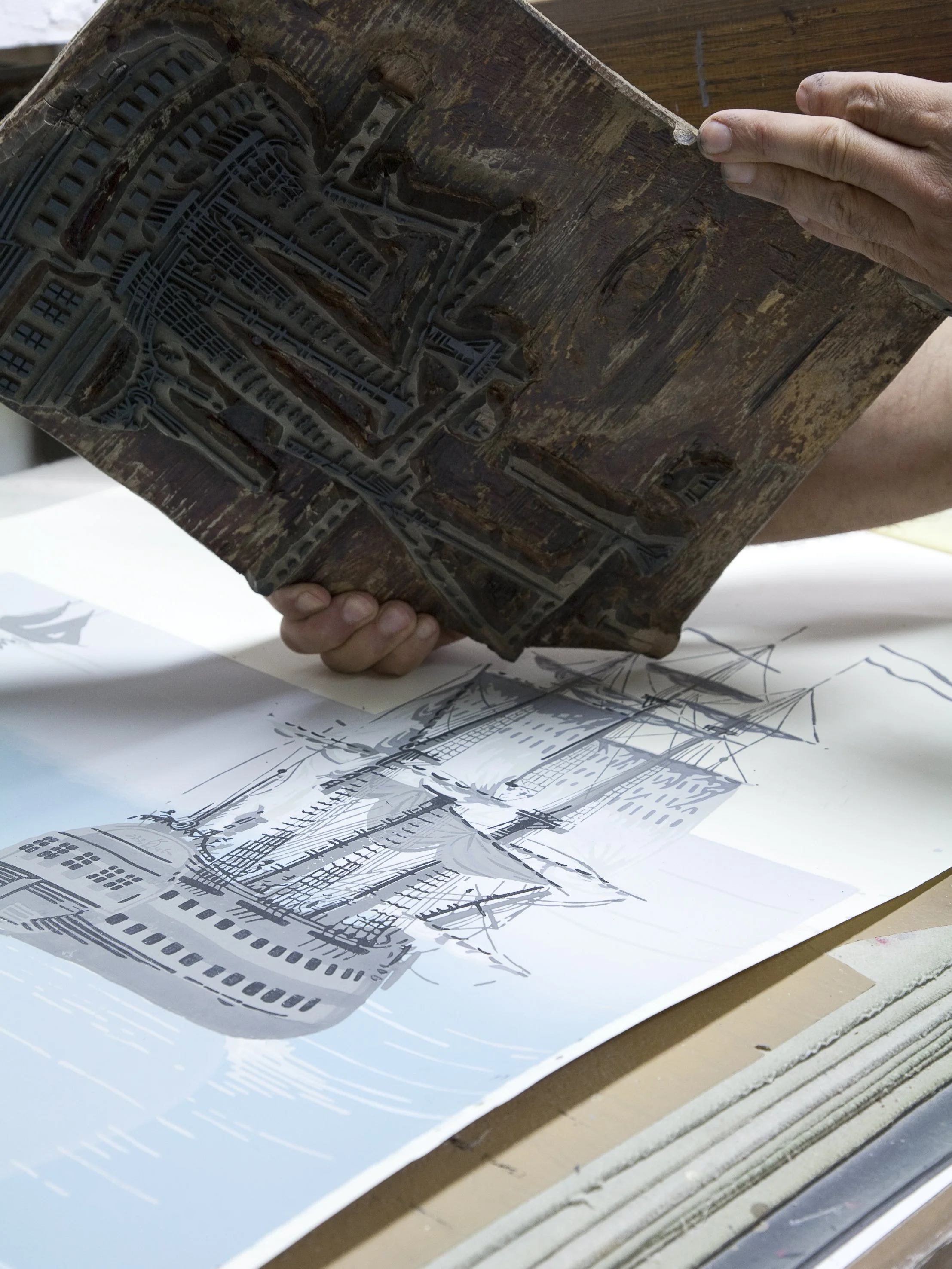 Person holding a wooden block with an intricate carved design over a large ship blueprint on a table.