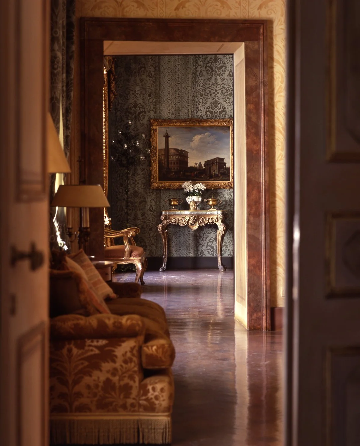 View of an elegant, vintage-style interior through a doorway, showing a sofa with pillows on the left, ornate side tables with lamps, a decorative console table with flowers, a large framed painting of a historic building, and patterned wallpaper.
