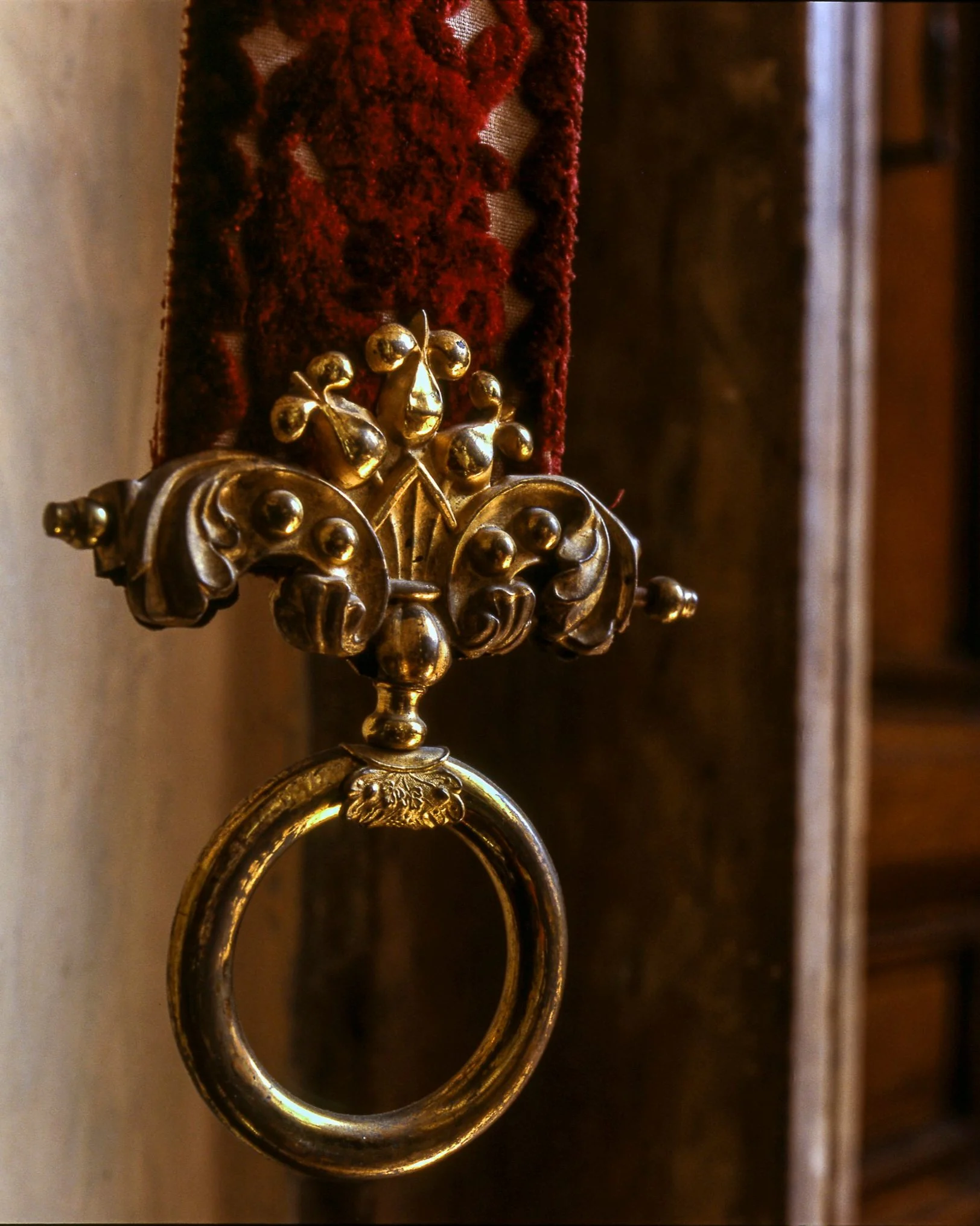 Close-up of an ornate, antique brass door knocker hanging from a red fabric strap against a wooden background.