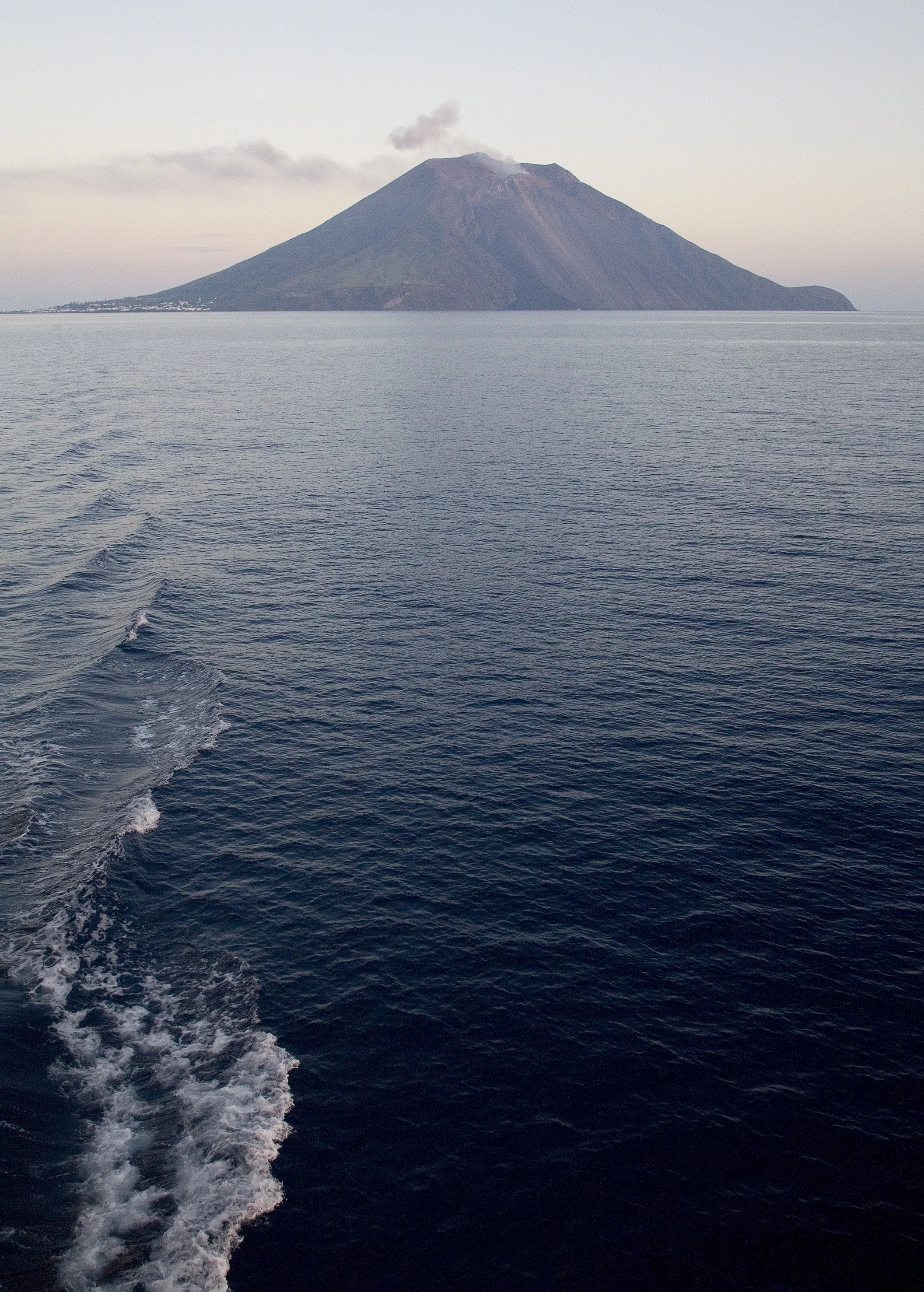 A view of a distant volcanic island with smoke rising from its summit, seen across a body of water with ripples, under a light sky.