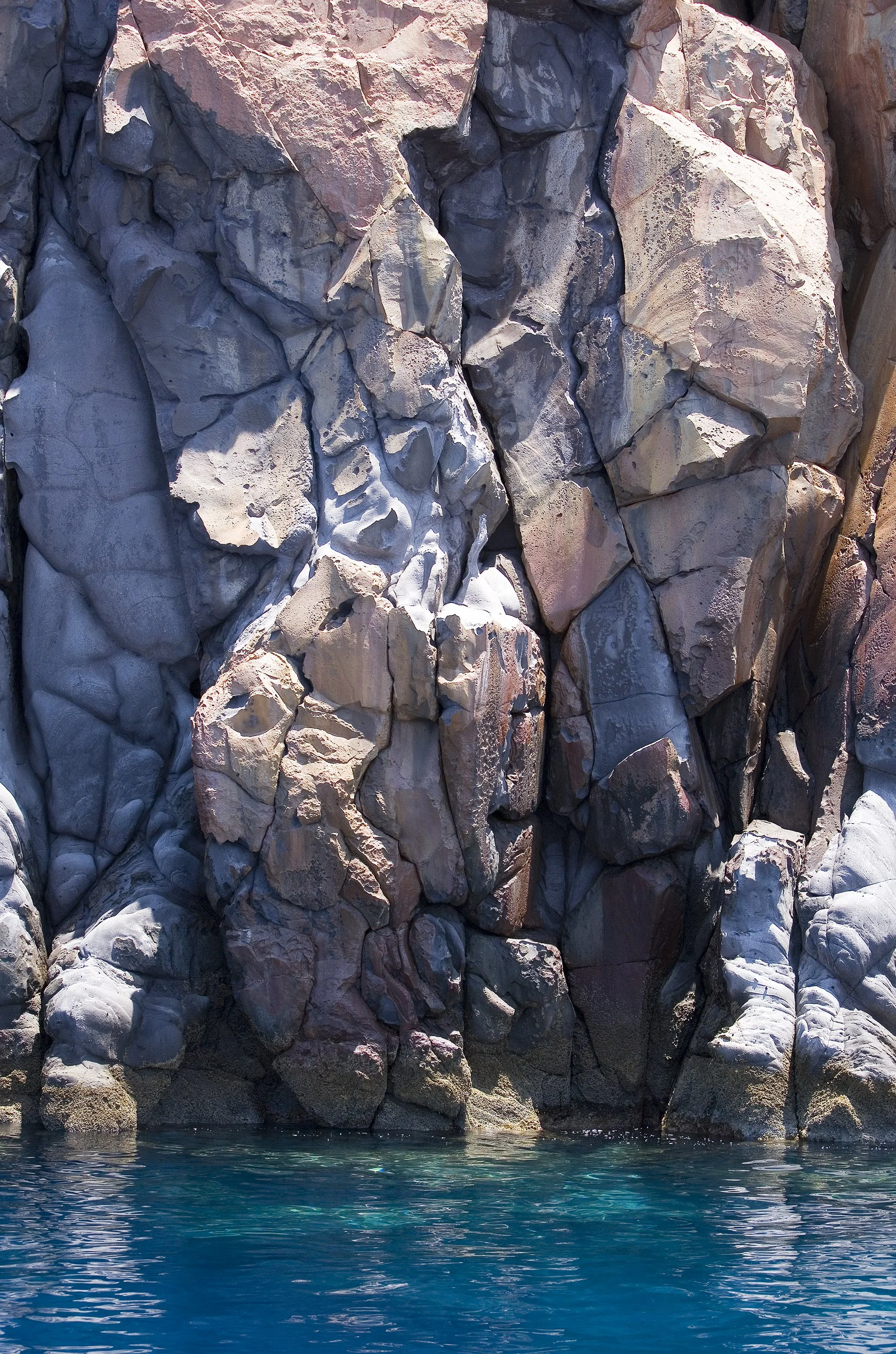 A large, rugged rock cliff with various shades of brown, gray, and pink, meeting clear blue water at the base.