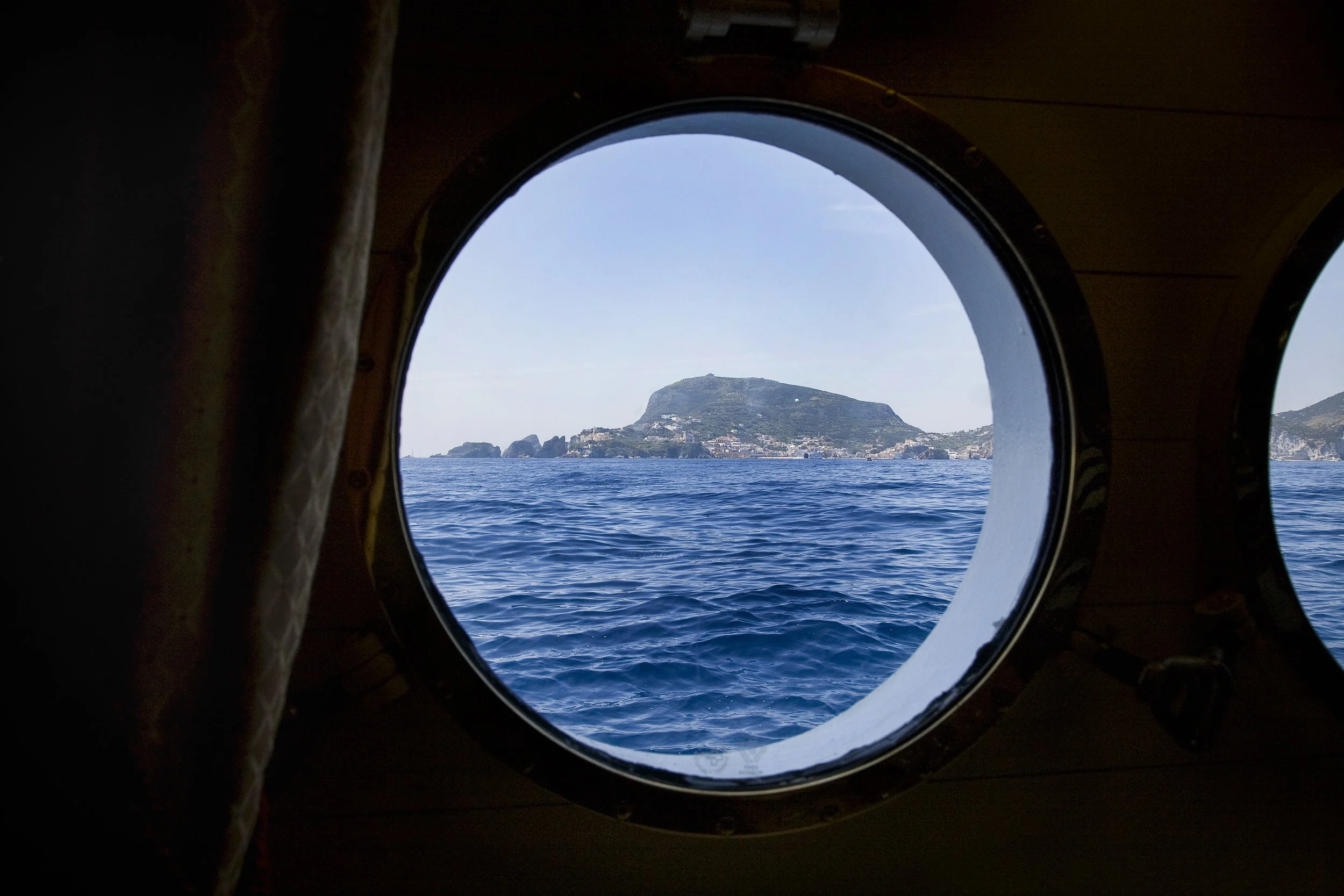 View of an island through a porthole on a boat, with blue ocean and sky.