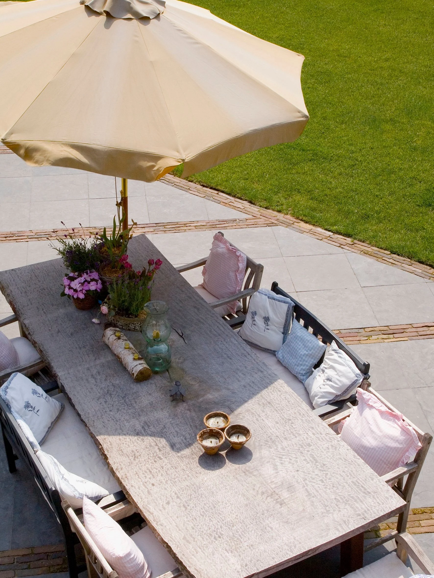 A large outdoor dining table with several throw pillows on the chairs, decorated with flowers, candles, and a large beige umbrella, set on a patio with a grassy yard nearby.