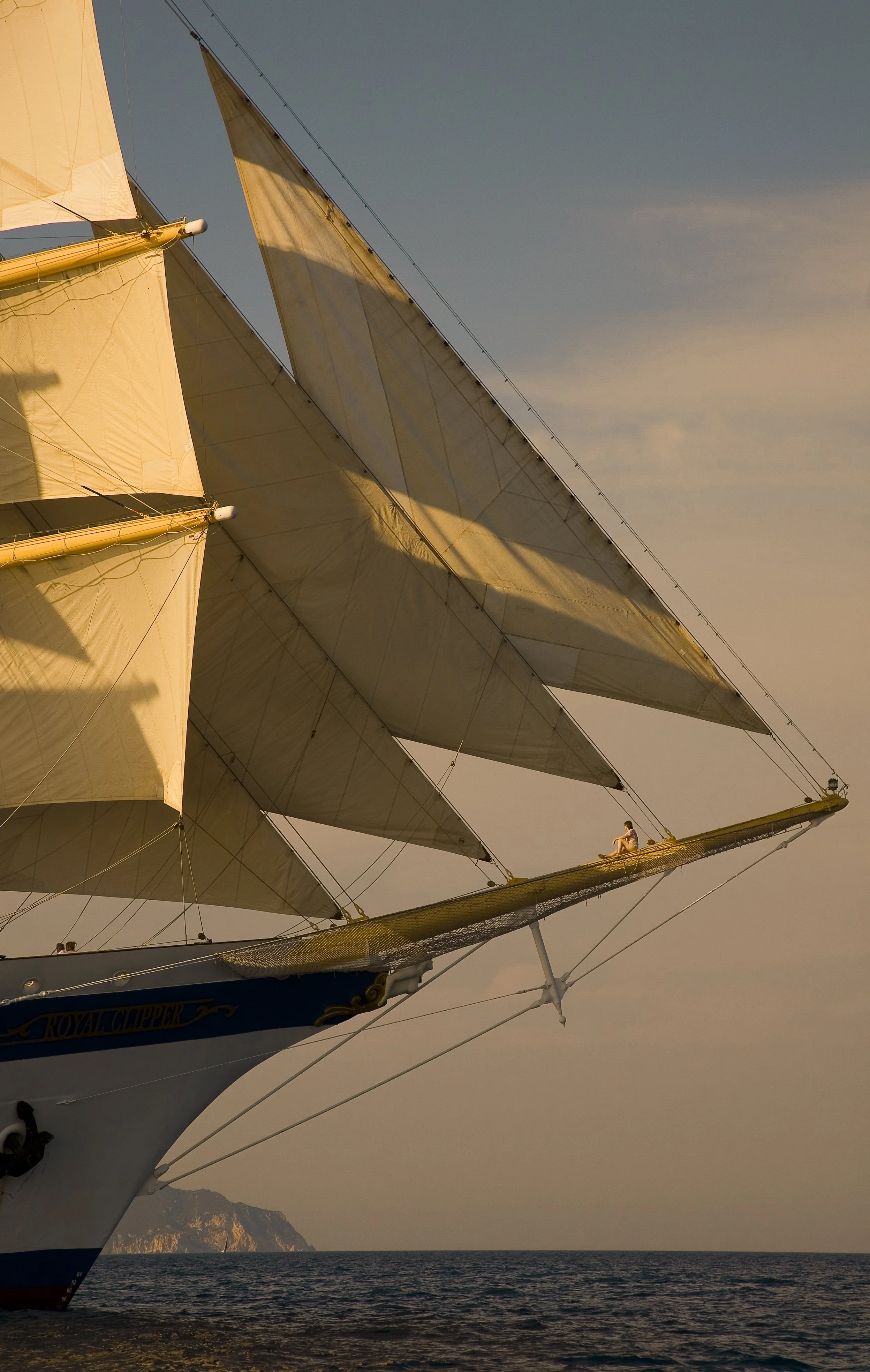 A large sailing ship with multiple sails is captured in this photograph. One person is sitting on the prow of the ship, enjoying the view. The sky is partly cloudy, and the ocean stretches to the horizon with a faint outline of a mountain in the distance.