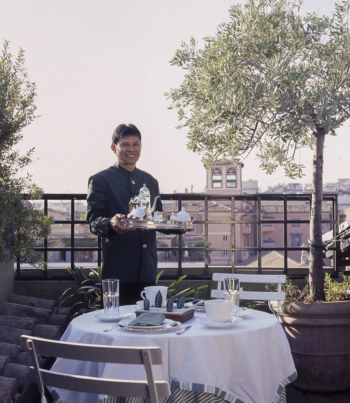 A man in a black traditional jacket holding a tray with a teapot and cups on a balcony, with a table set for tea in front of him. There are plants, trees, and a cityscape in the background.