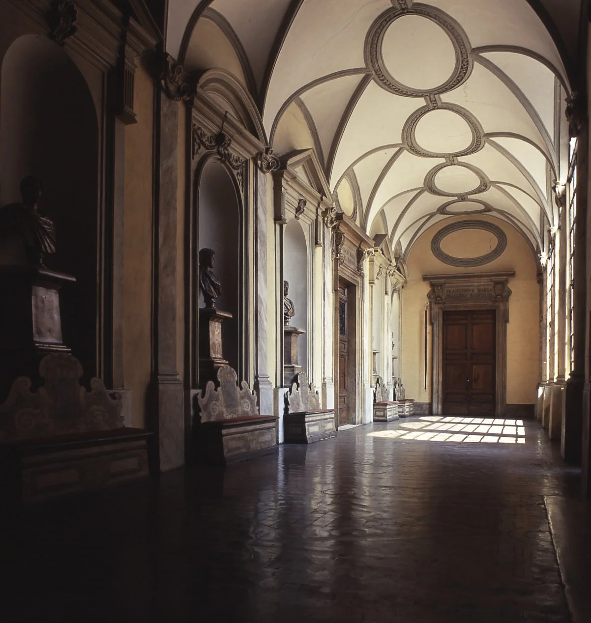 A grand hallway with arched ceilings, decorative moldings, bust sculptures on pedestals, and a wooden door at the end, illuminated by natural light coming through windows.