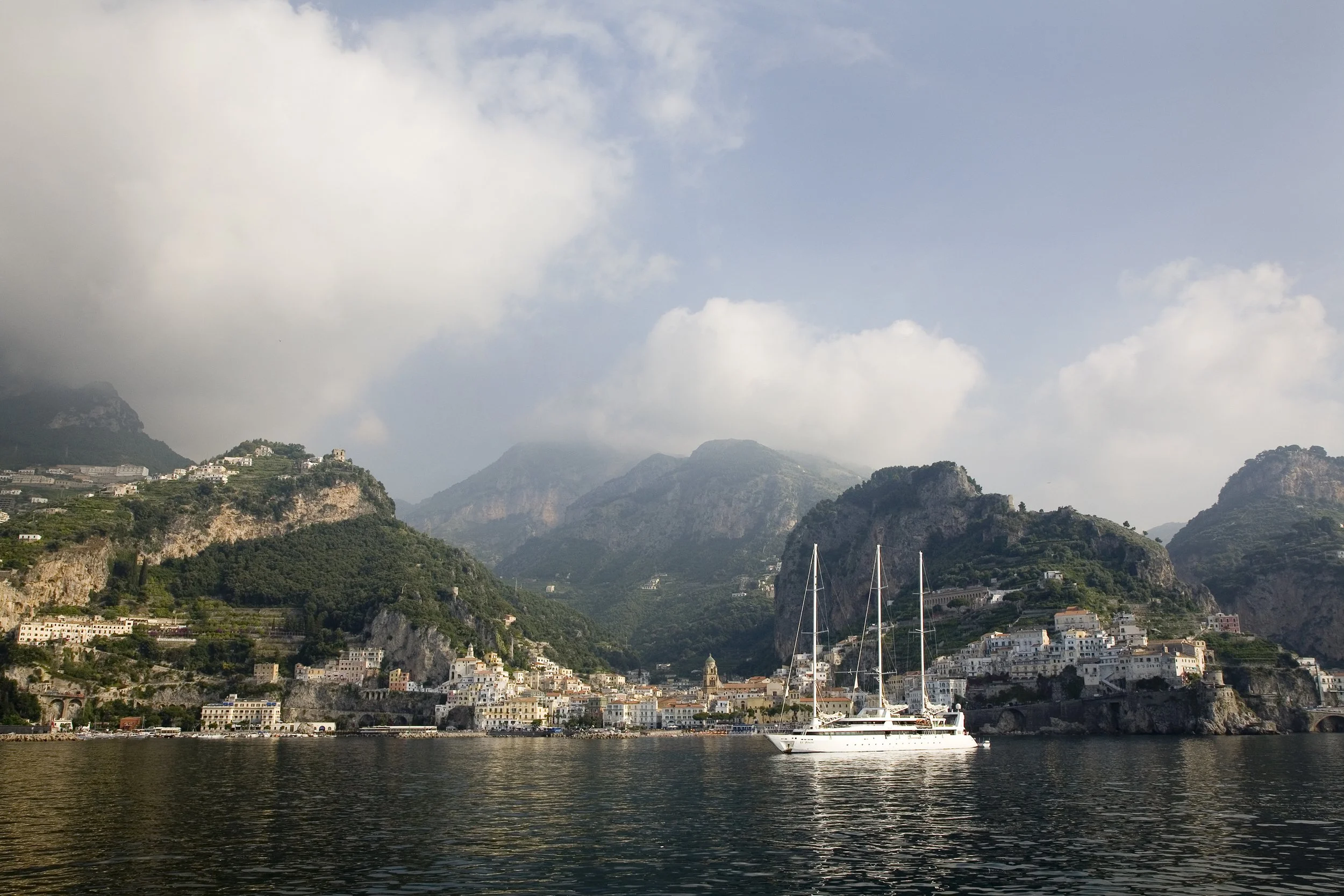 Coastal town with white buildings on hilly terrain, mountains in background, sailboat on calm water in the foreground, partly cloudy sky.