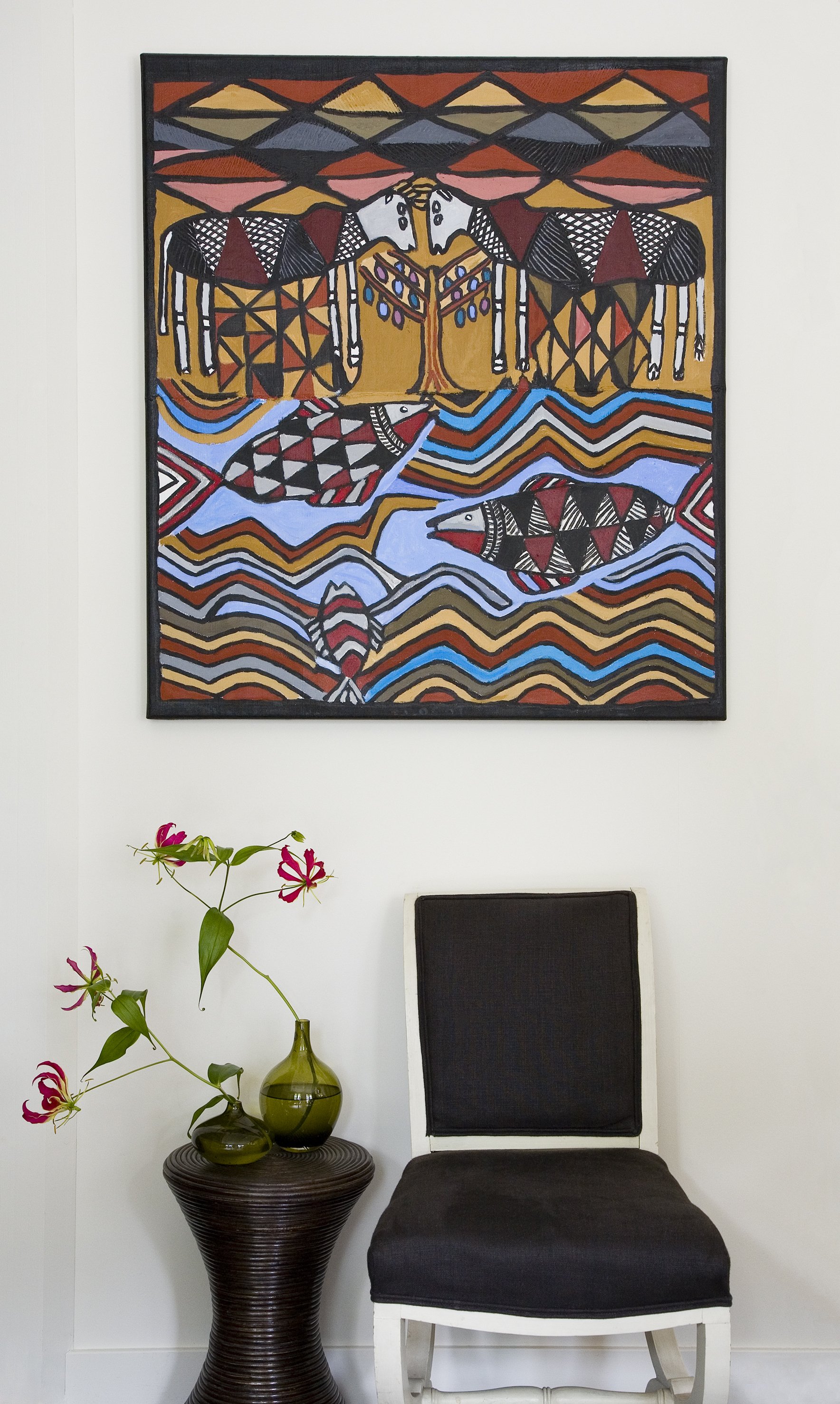 A wall decor piece with an abstract tribal art design, featuring animals, fish, water, and geometric patterns in red, blue, black, beige, and yellow. A black chair and a dark wooden side table with two green vases holding pink and red flowers are beneath the art.