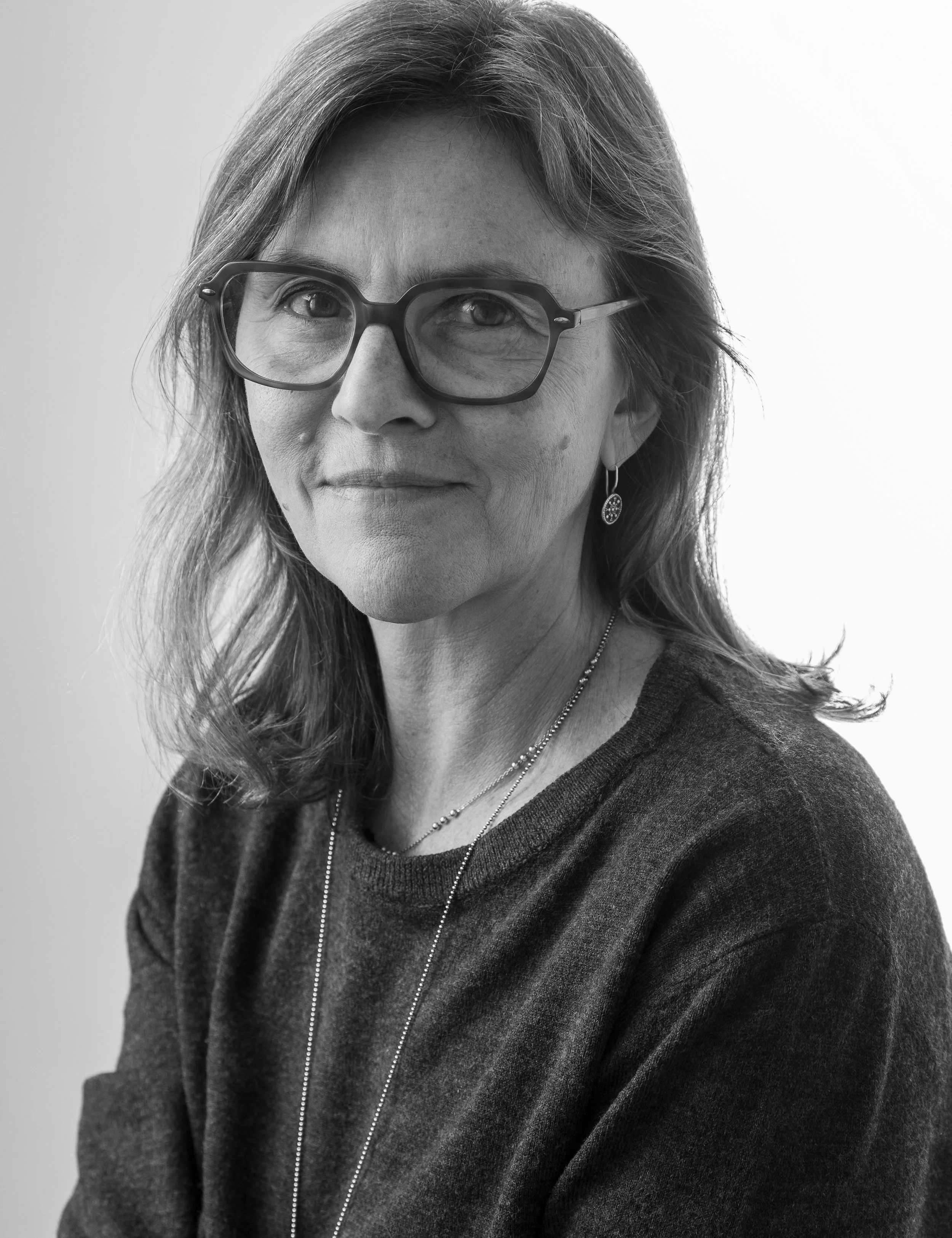 A black and white portrait of a middle-aged woman with glasses, wearing a dark sweater, earrings, and a layered necklace, looking at the camera with a slight smile.