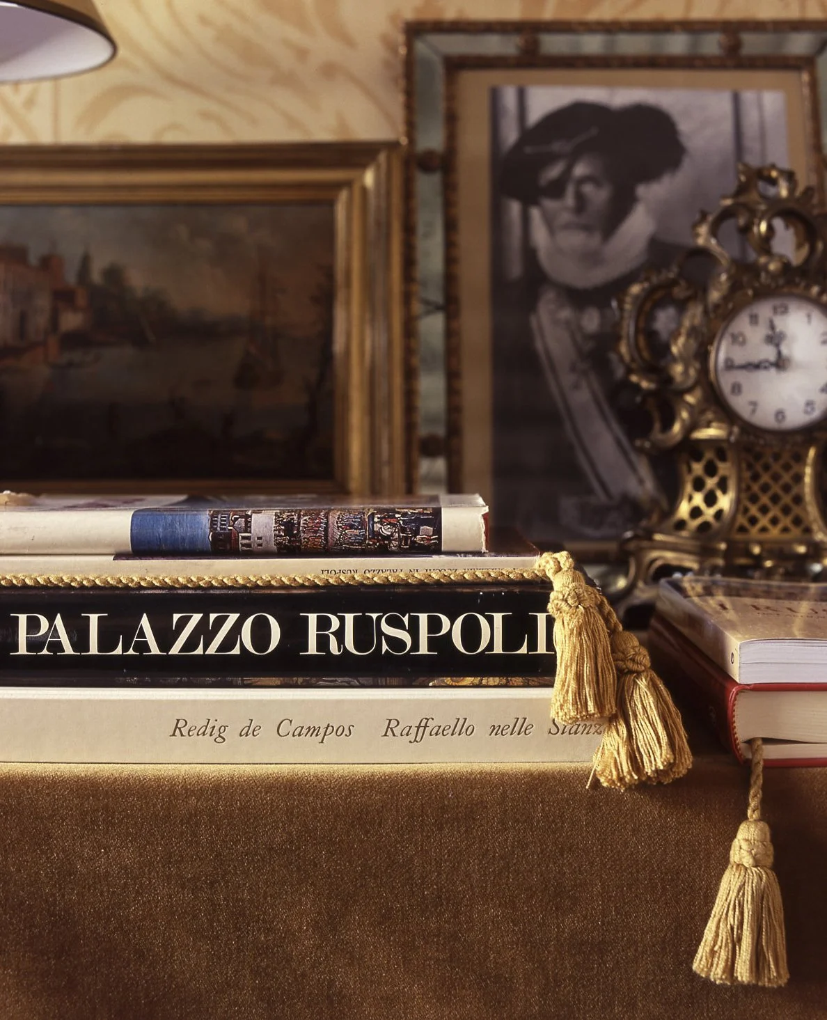Close-up of a stack of books on a table with vintage decor in the background, including framed black-and-white photographs and an ornate clock.