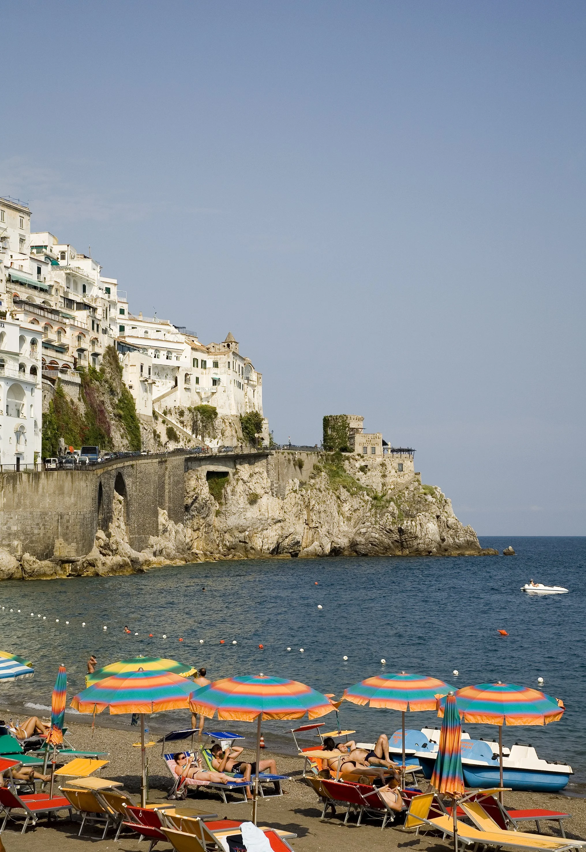 Beach with colorful umbrellas and sunbathers, overlooking rocky cliffs with white buildings on a seaside.