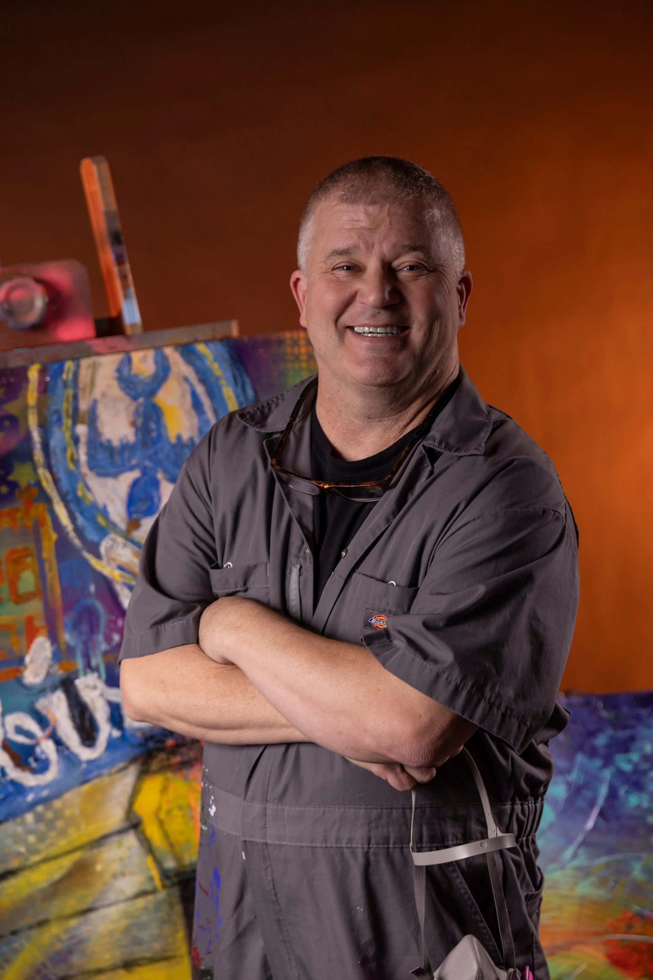 A man with short gray hair, wearing a gray work jacket with his arms crossed, smiling in front of an abstract colorful painted backdrop.