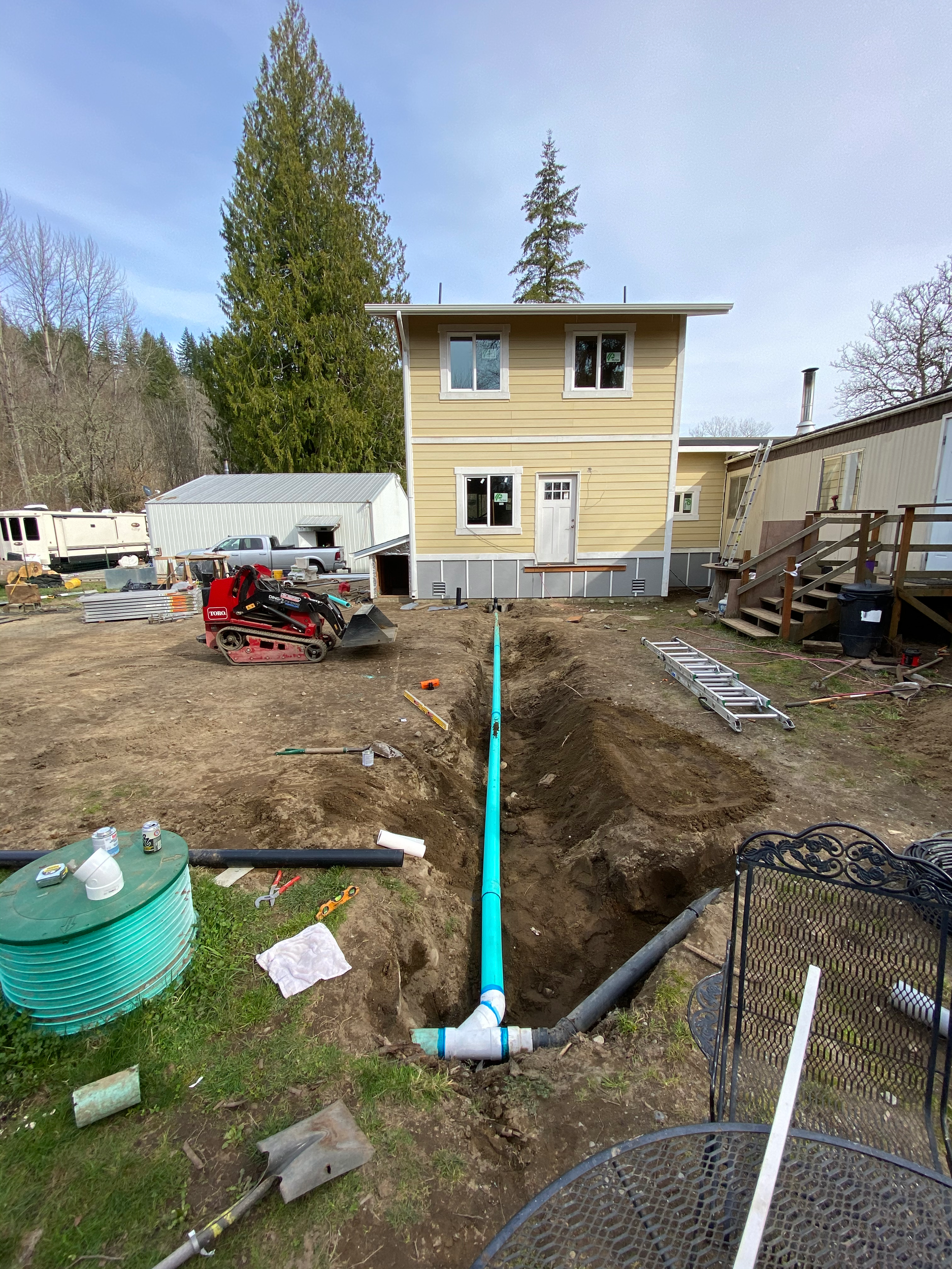 An ongoing construction or plumbing project outside a yellow house with white trim. A section of the ground is dug up, with a blue pipe and other tools laying nearby, indicating pipe installation or repair work.