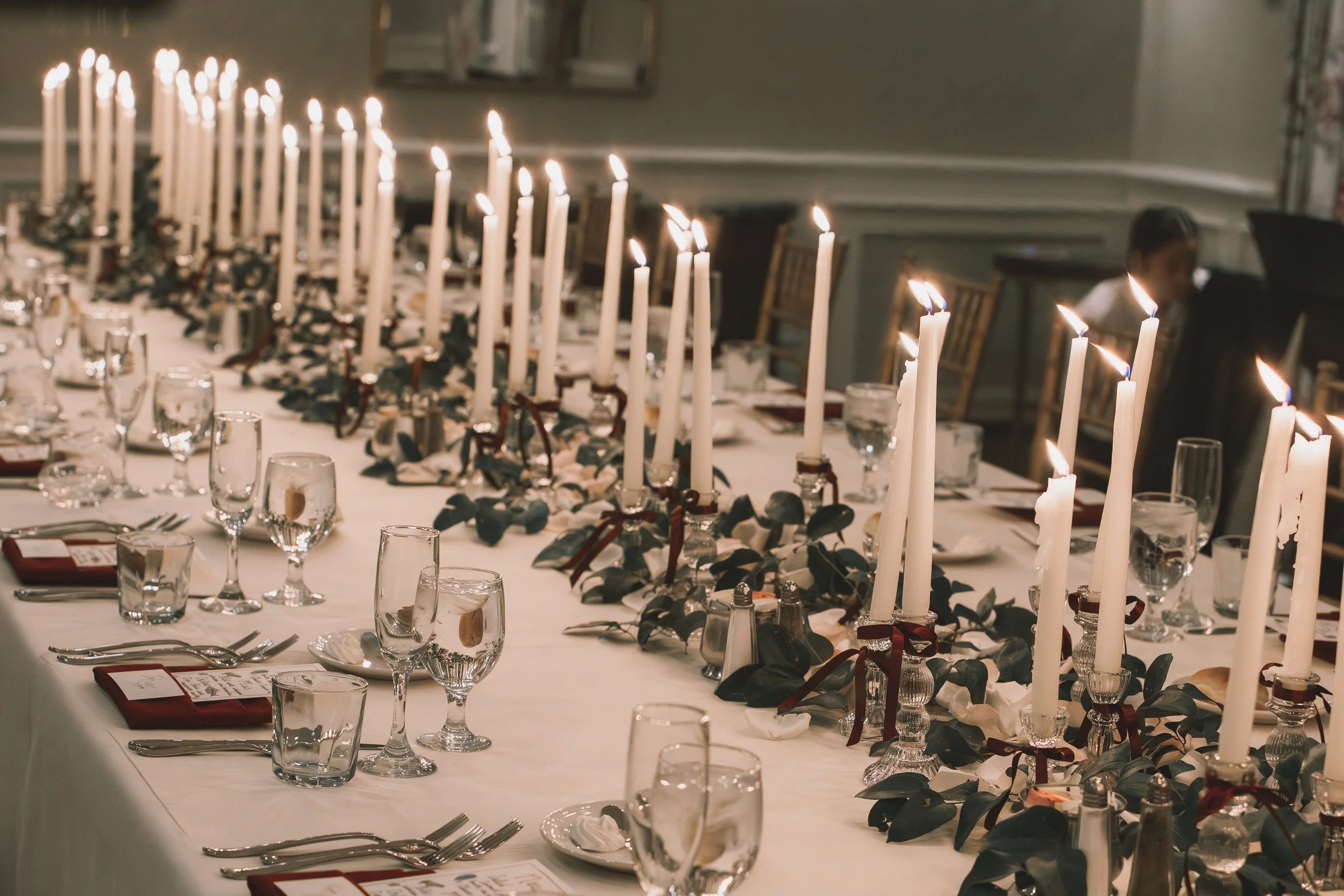 A long dining table decorated with multiple white candles in candle holders, surrounded by greenery and candles in small glasses, set for a formal dinner with glasses, silverware, and red napkins.
