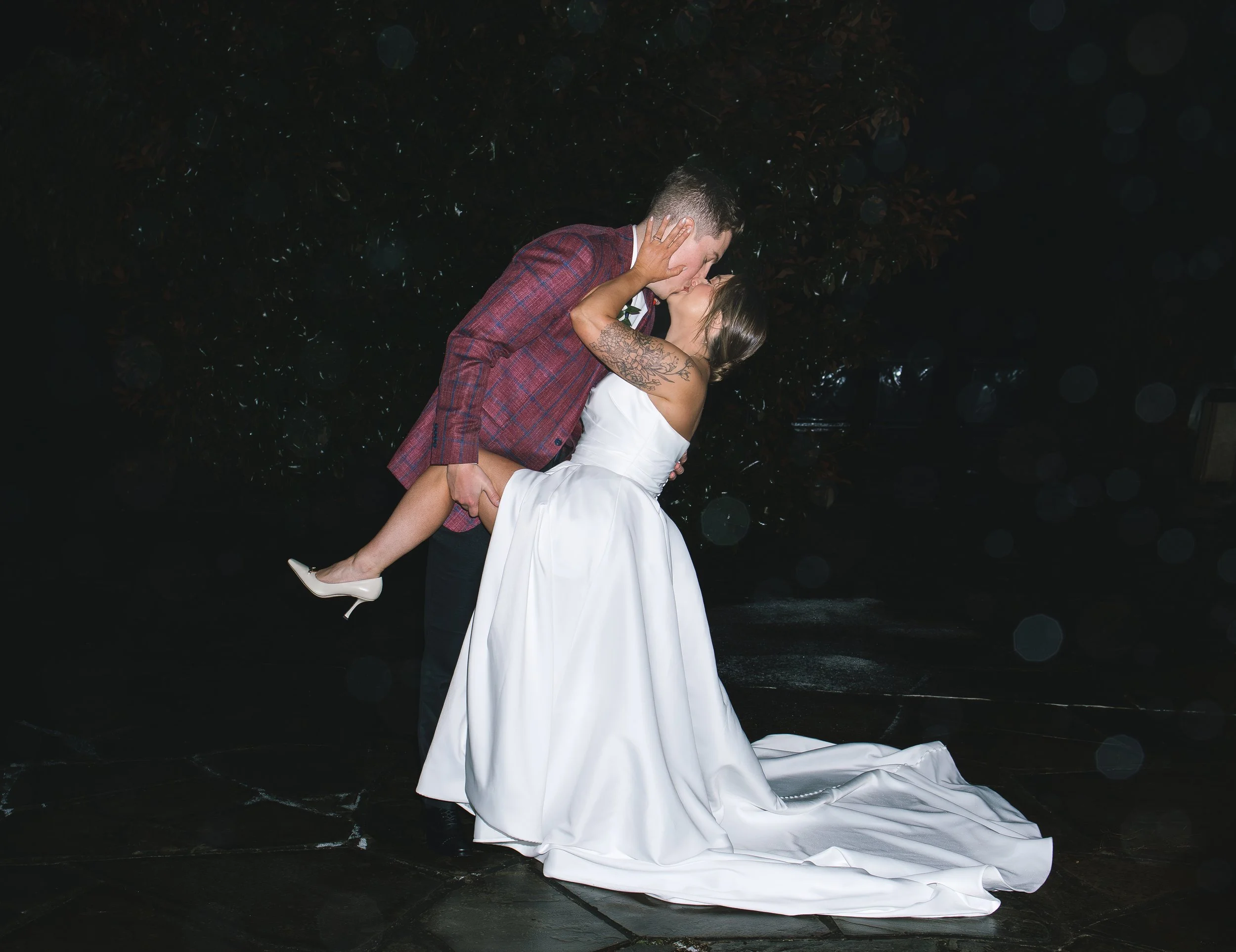 Couple kissing outdoors at night, the man lifting the woman in a white wedding gown, wearing beige heels, dark background with trees.