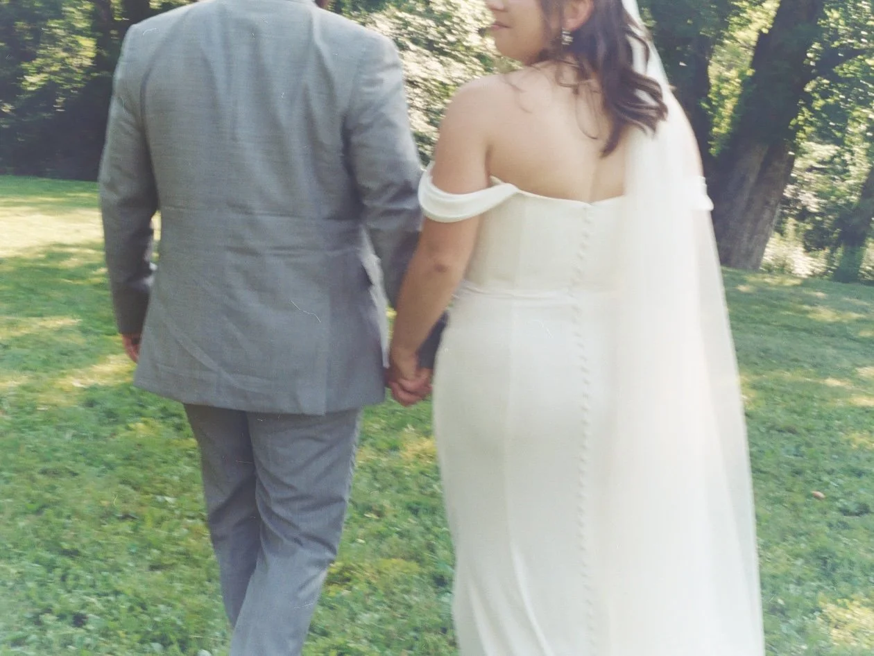 A bride and groom holding hands in a park, seen from behind, during their wedding. The bride wears an off-the-shoulder white wedding dress, and the groom wears a gray suit.