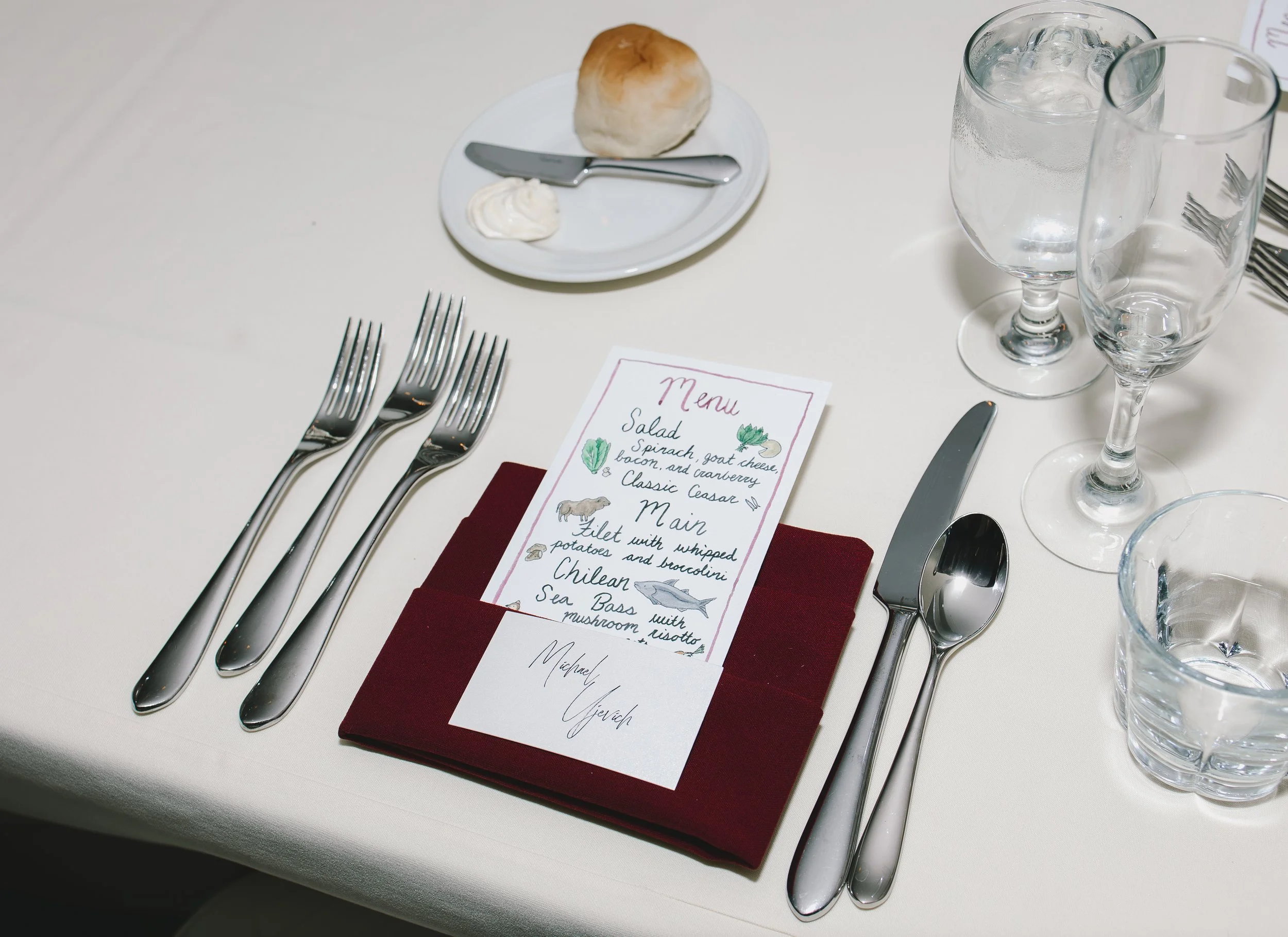 A formal table setting with silverware, water glasses, a folded burgundy napkin, and a menu card on top. There is also a small plate with a bread roll, knife, and a dollop of butter.