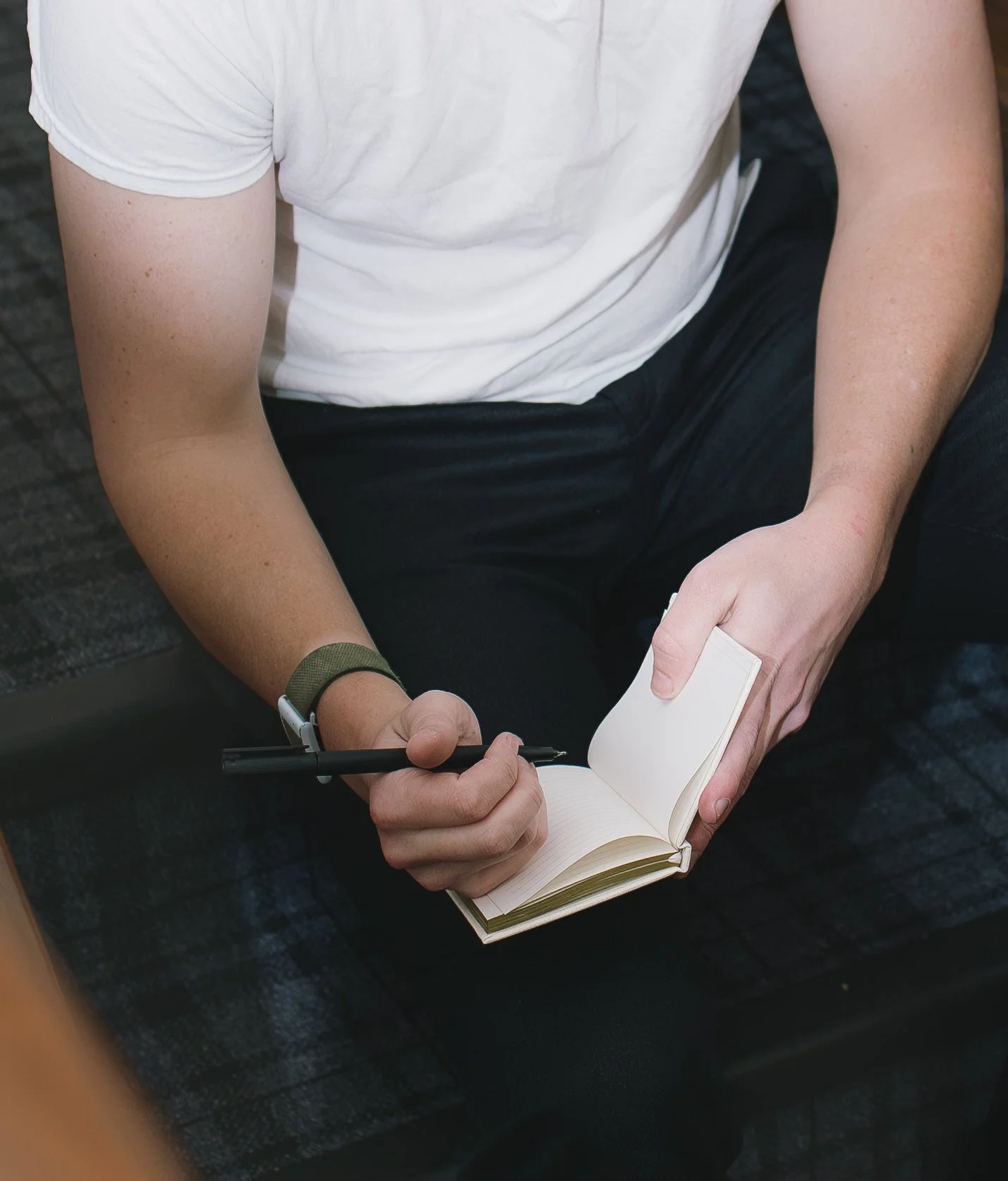 A person wearing a white T-shirt and black pants sitting on a black chair, holding an open blank notebook and a black pen.