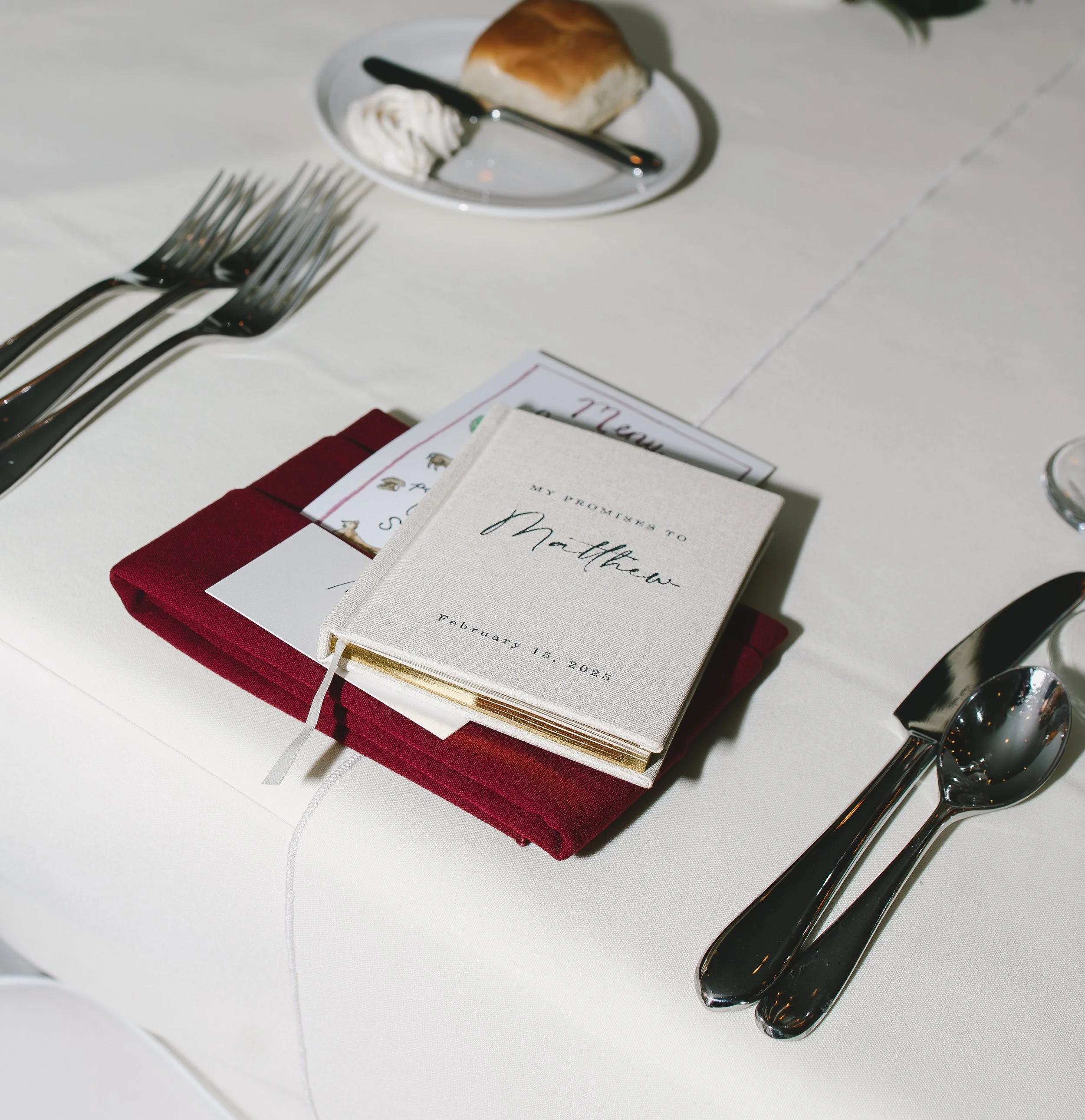 Wedding place setting with a white tablecloth, silverware, a burgundy napkin, a guest book with the name Matthew and the date February 15, 2026, and a small plate with a bread roll and butter.