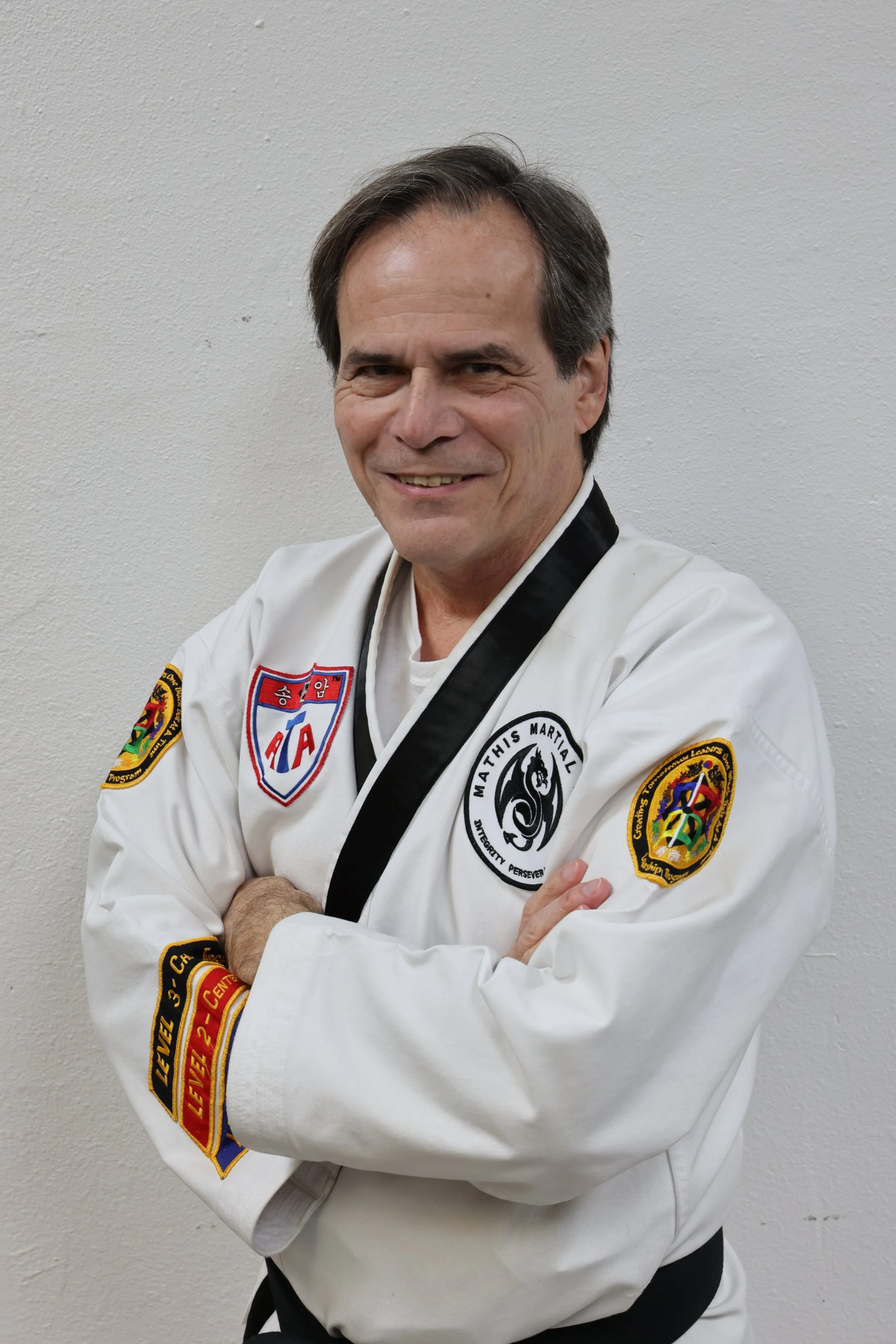 A middle-aged man with gray hair and a mustache, smiling, wearing a black polo shirt with a logo, standing with arms crossed against a red background.