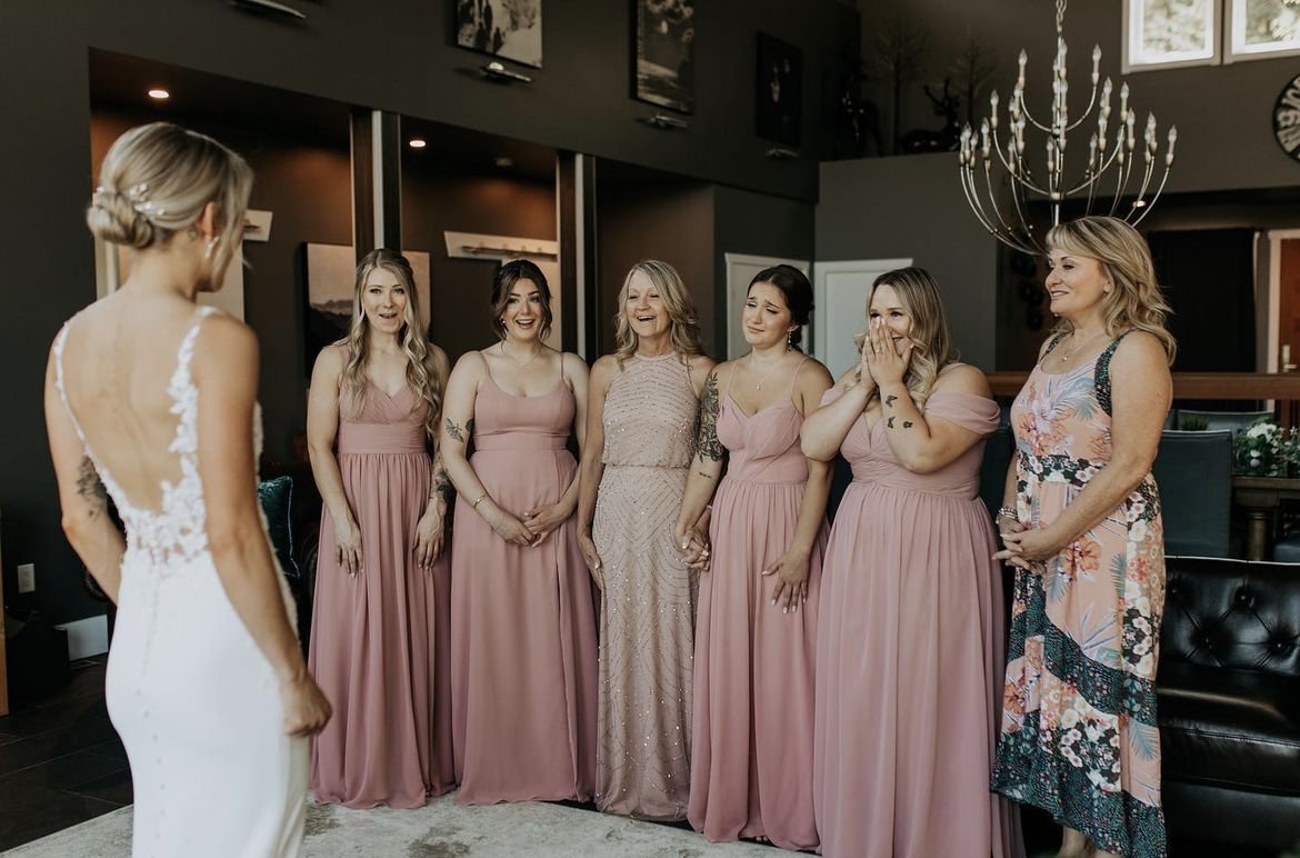 Bride in white dress with group of bridesmaids and women in pink dresses, reacting emotionally indoors, chandelier visible.