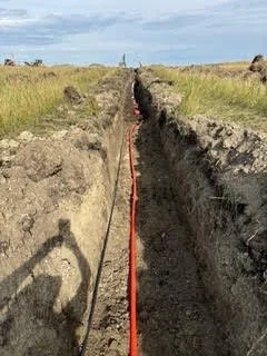 A construction or excavation site with a long, narrow trench in the ground, surrounded by grassy fields, and a red line marking the trench's path.