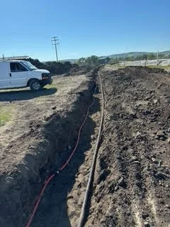 Construction site with a trench, utility pipes, and a white van in a rural area with hills in the background.